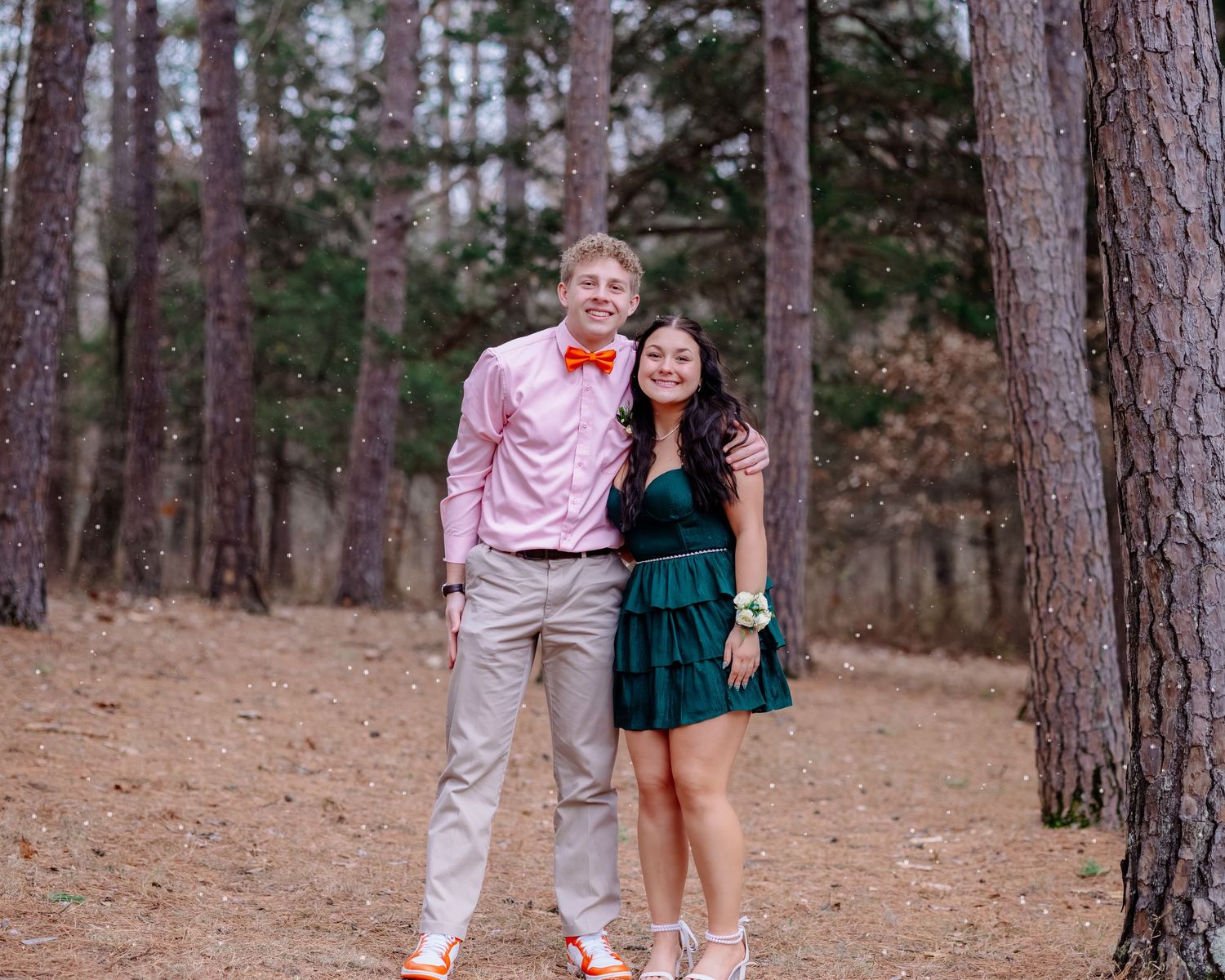 A couple poses together in a pine forest, with one wearing a pink shirt and bowtie and the other in a green dress.