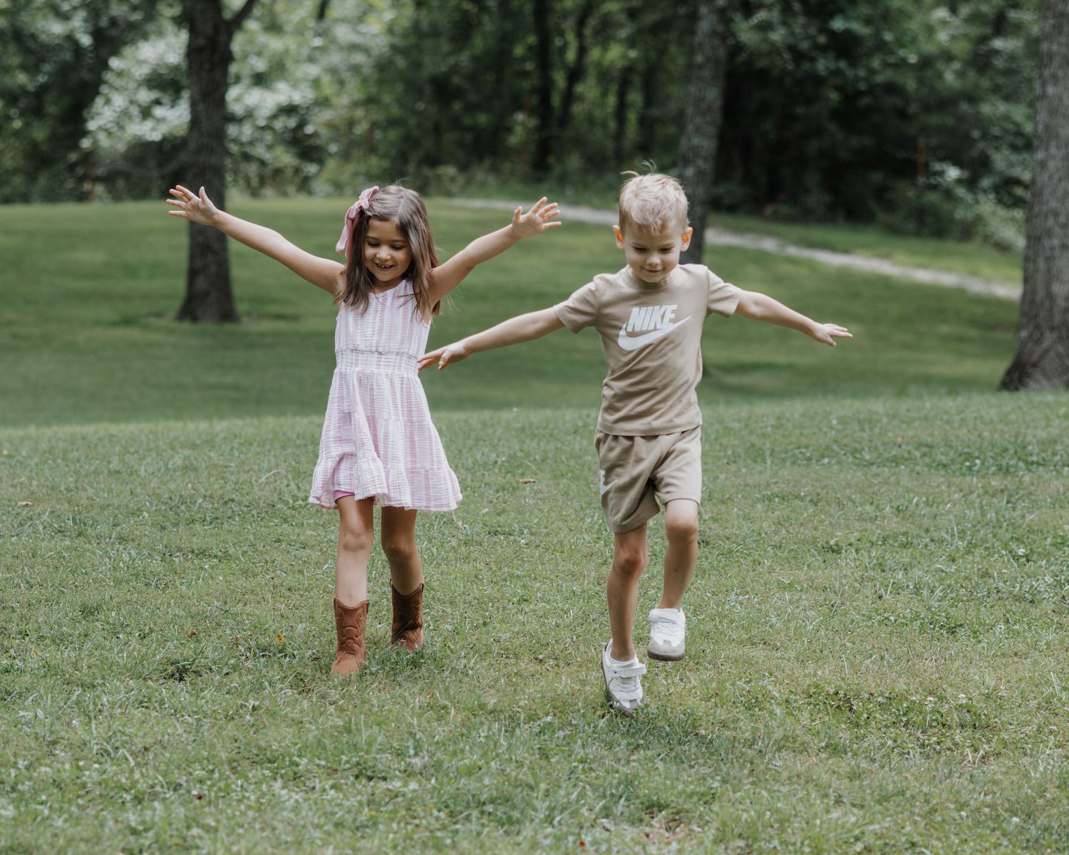 Two children play and jump together with outstretched arms on a grassy lawn surrounded by trees.