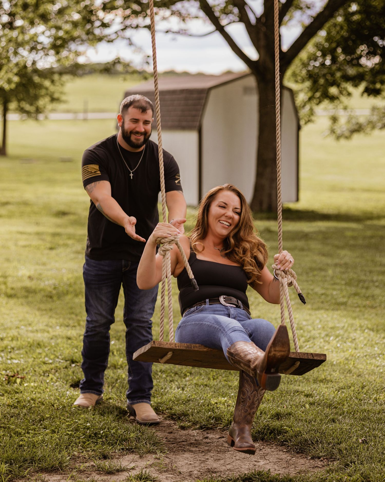 Man pushing smiling woman on rustic wooden rope swing in lush green backyard near white shed.