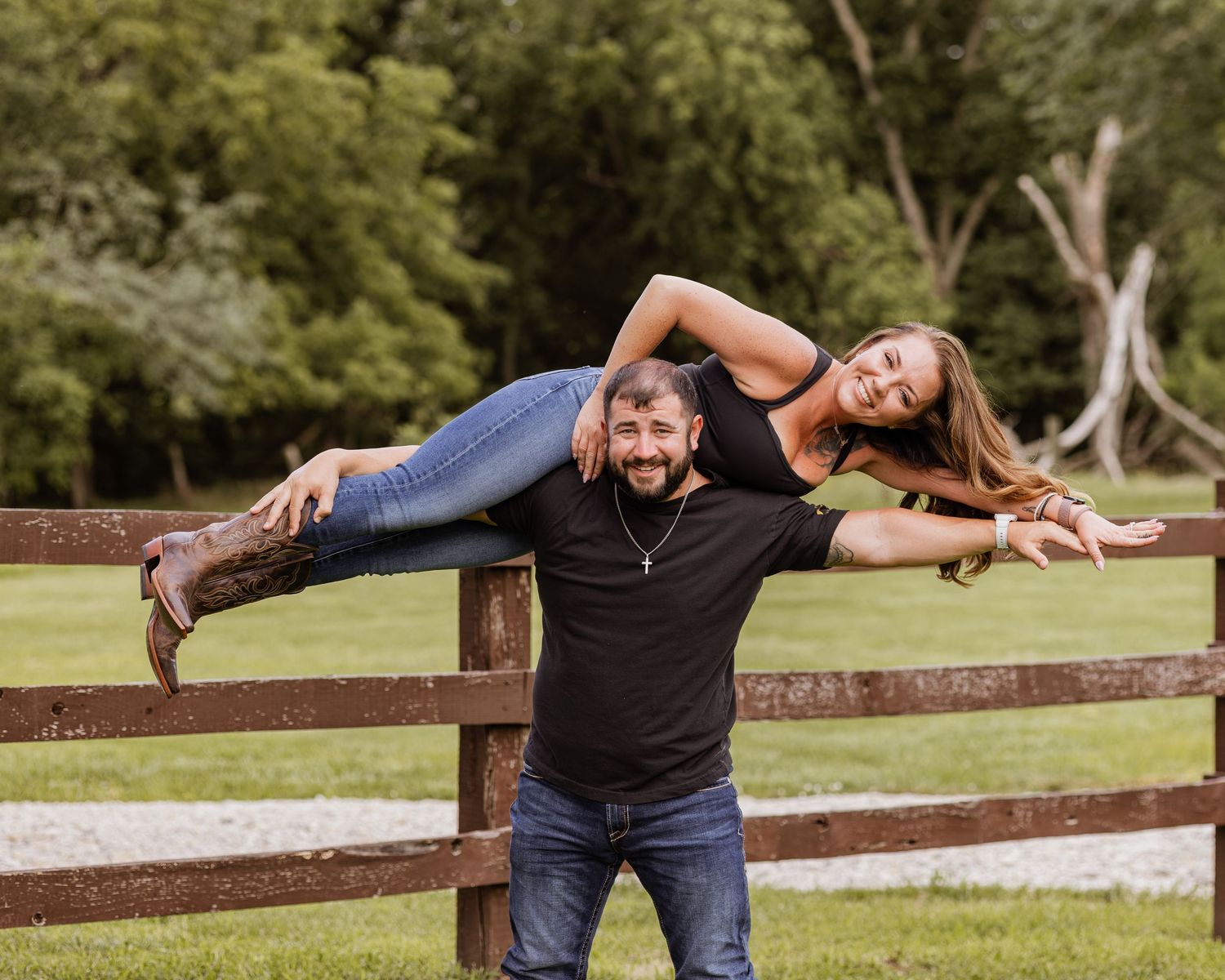 Man playfully lifting woman over his shoulder near a wooden fence on a green farm property.