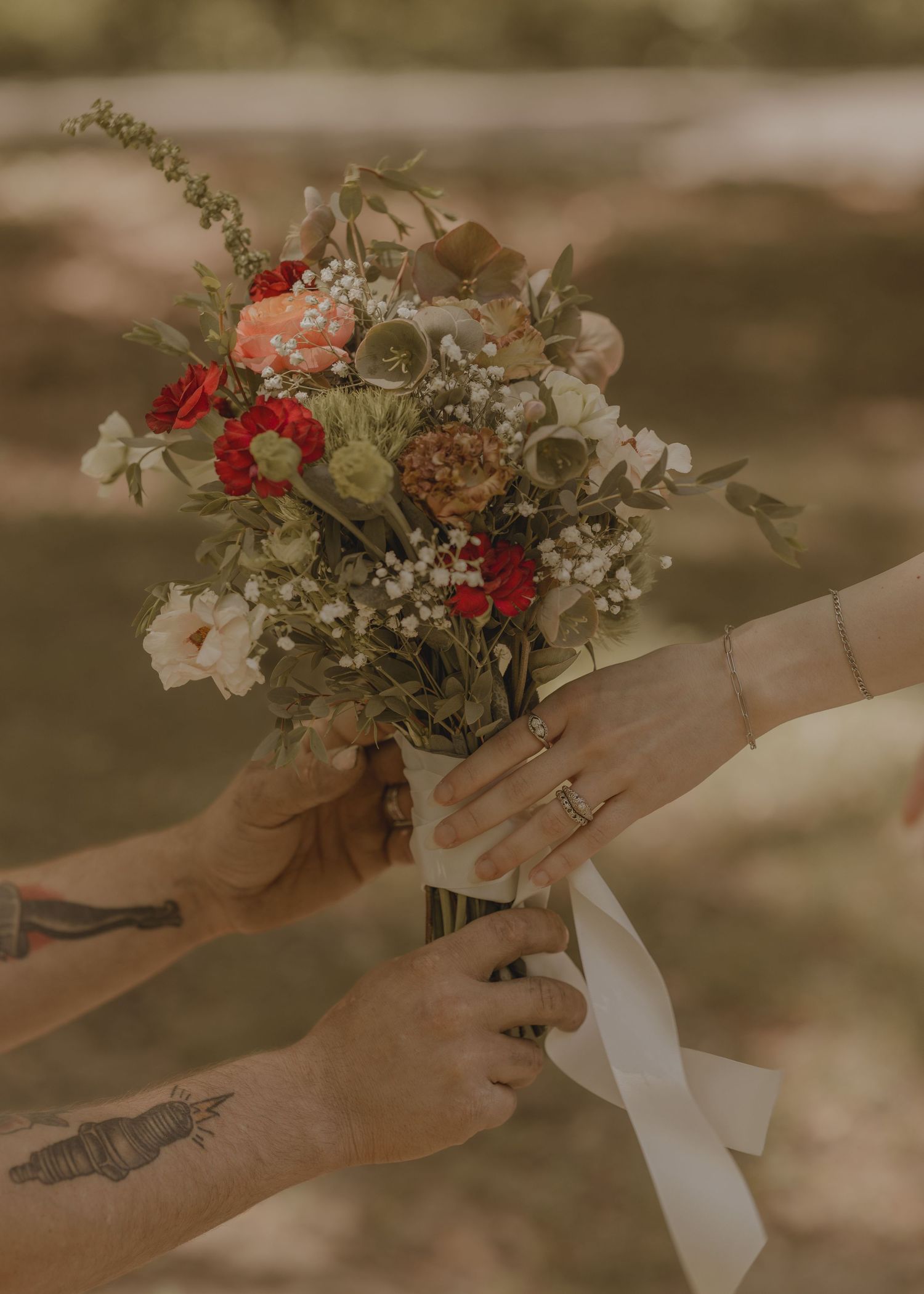 Couple exchanging a bridal bouquet of red, peach, and white flowers tied with a white ribbon.