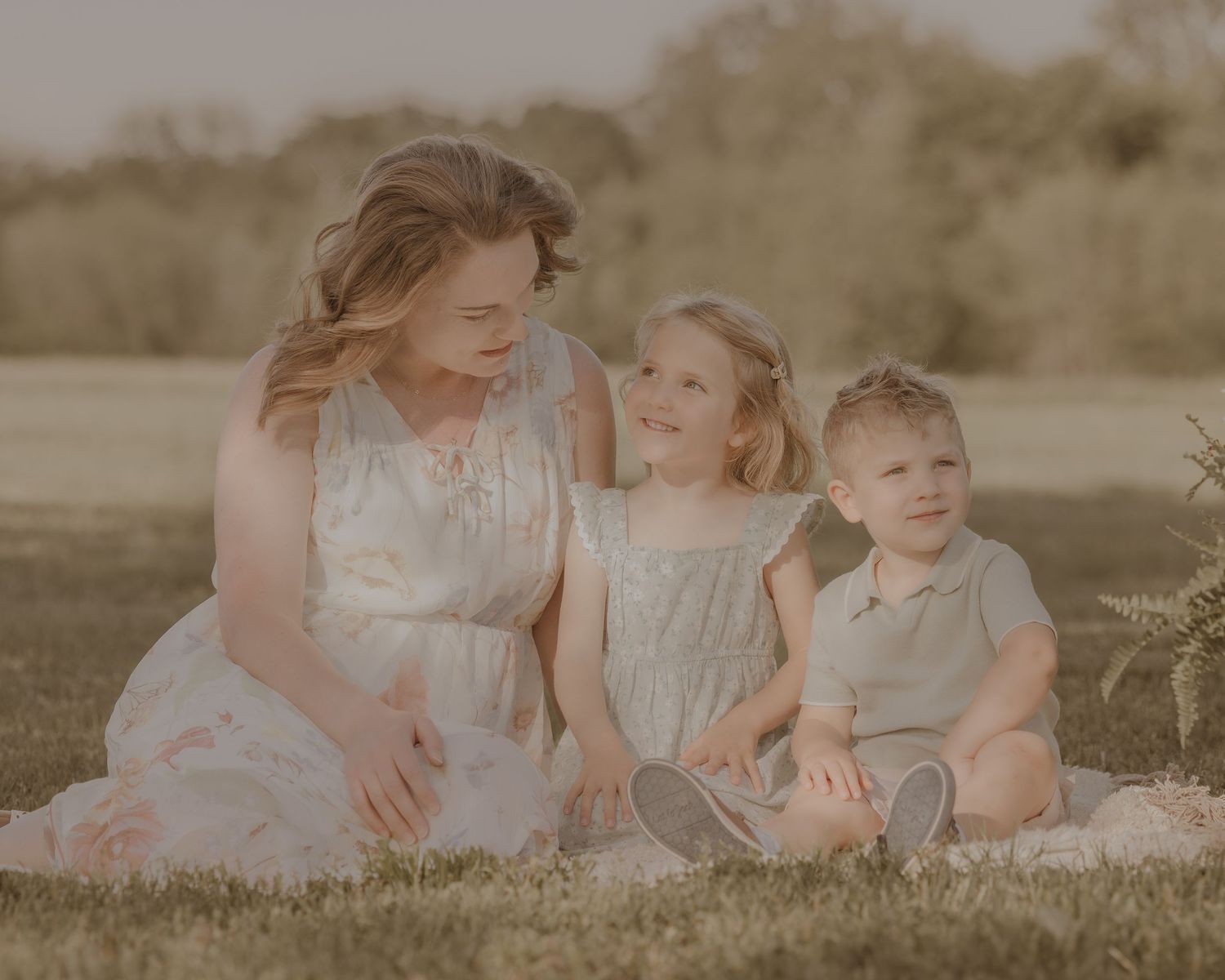 Woman in floral dress sitting outdoors with smiling girl and boy during warm sunset family portrait session.