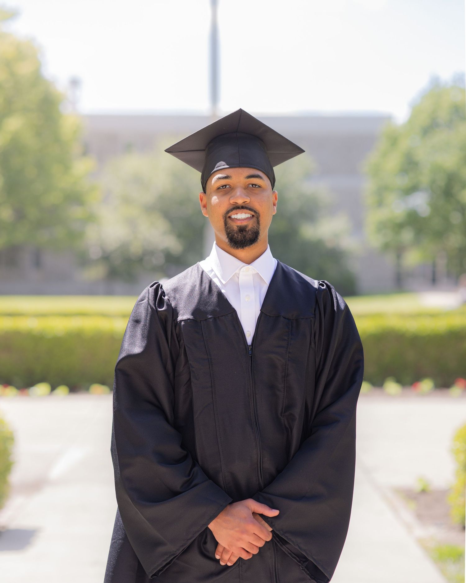 Smiling graduate in black cap and gown posing outdoors on a sunny campus.