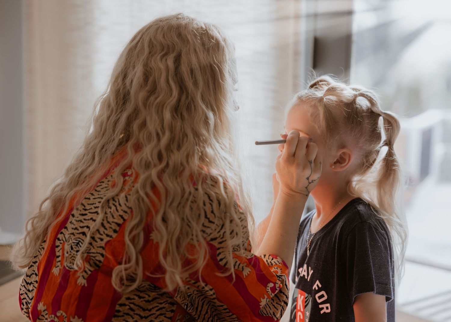 A woman with curly blonde hair applies makeup to a young girl near a bright window indoors.