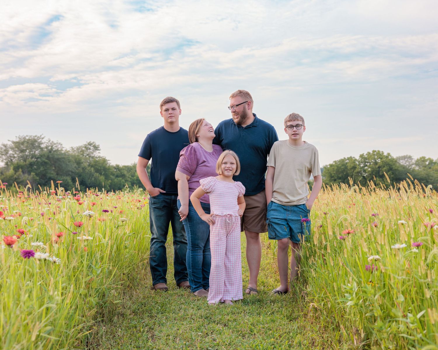 Family of five posing in a wildflower meadow path under a cloudy sky during golden hour.