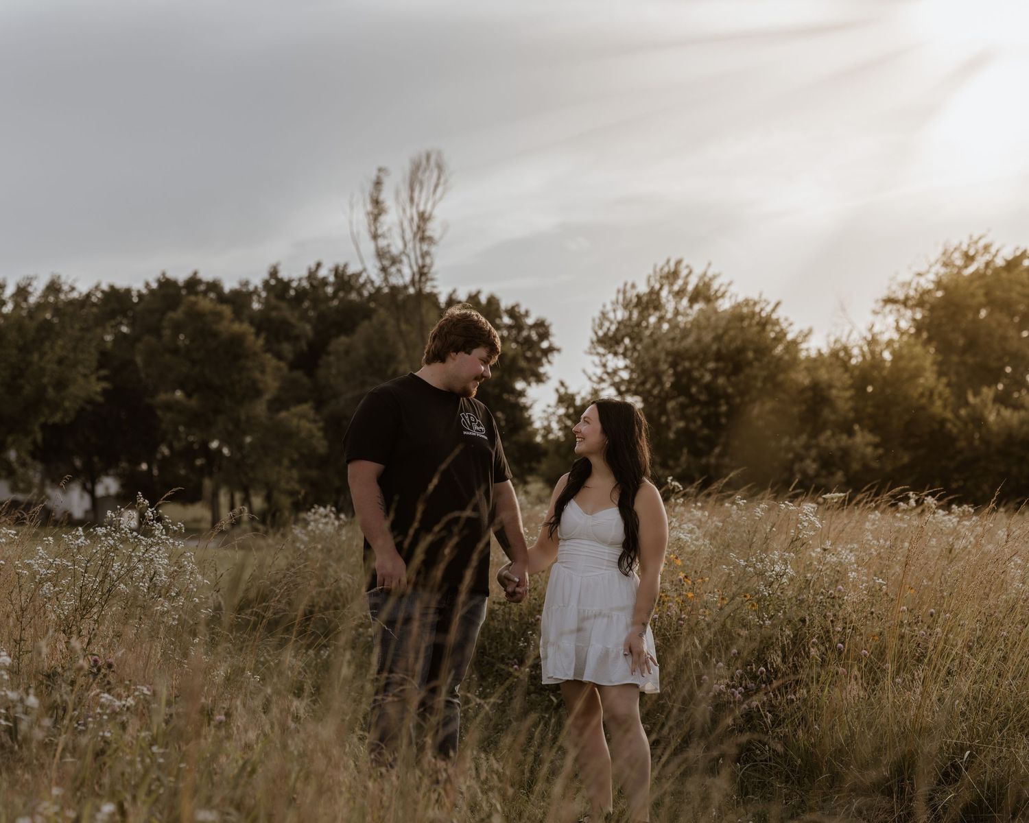 Couple standing in a golden field of tall grass at sunset, gazing into each other's eyes.