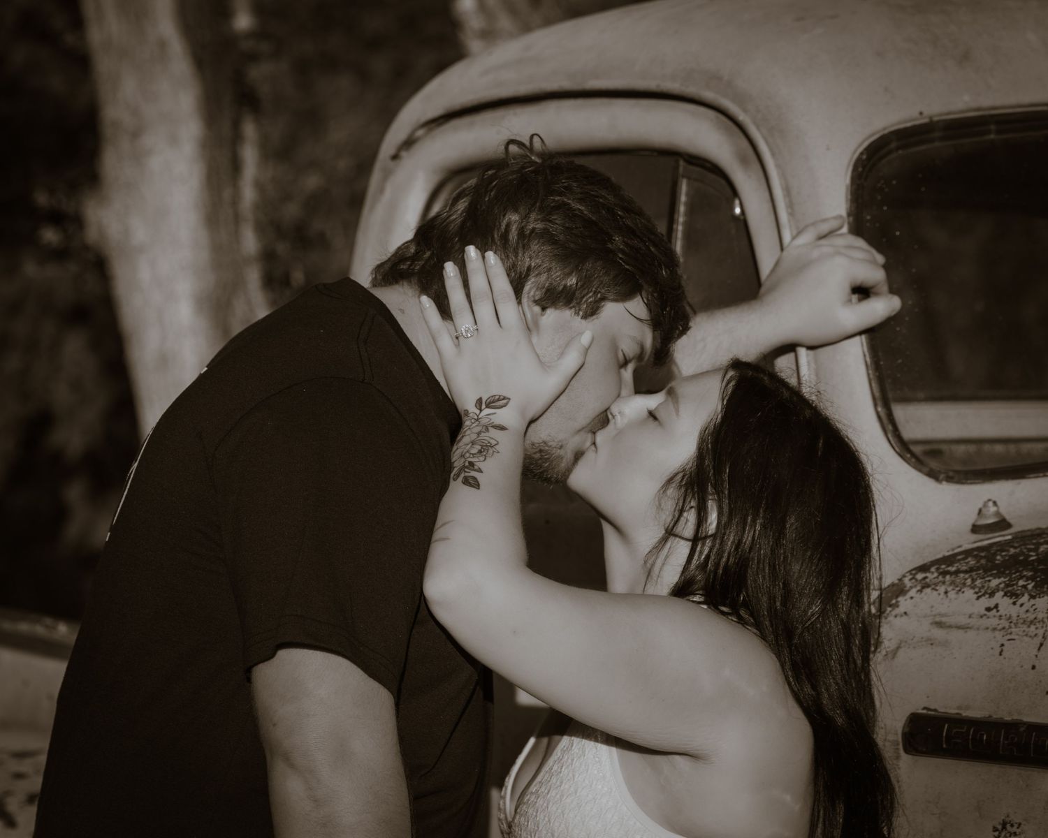 Couple sharing romantic kiss leaning against vintage truck in sepia toned photo.