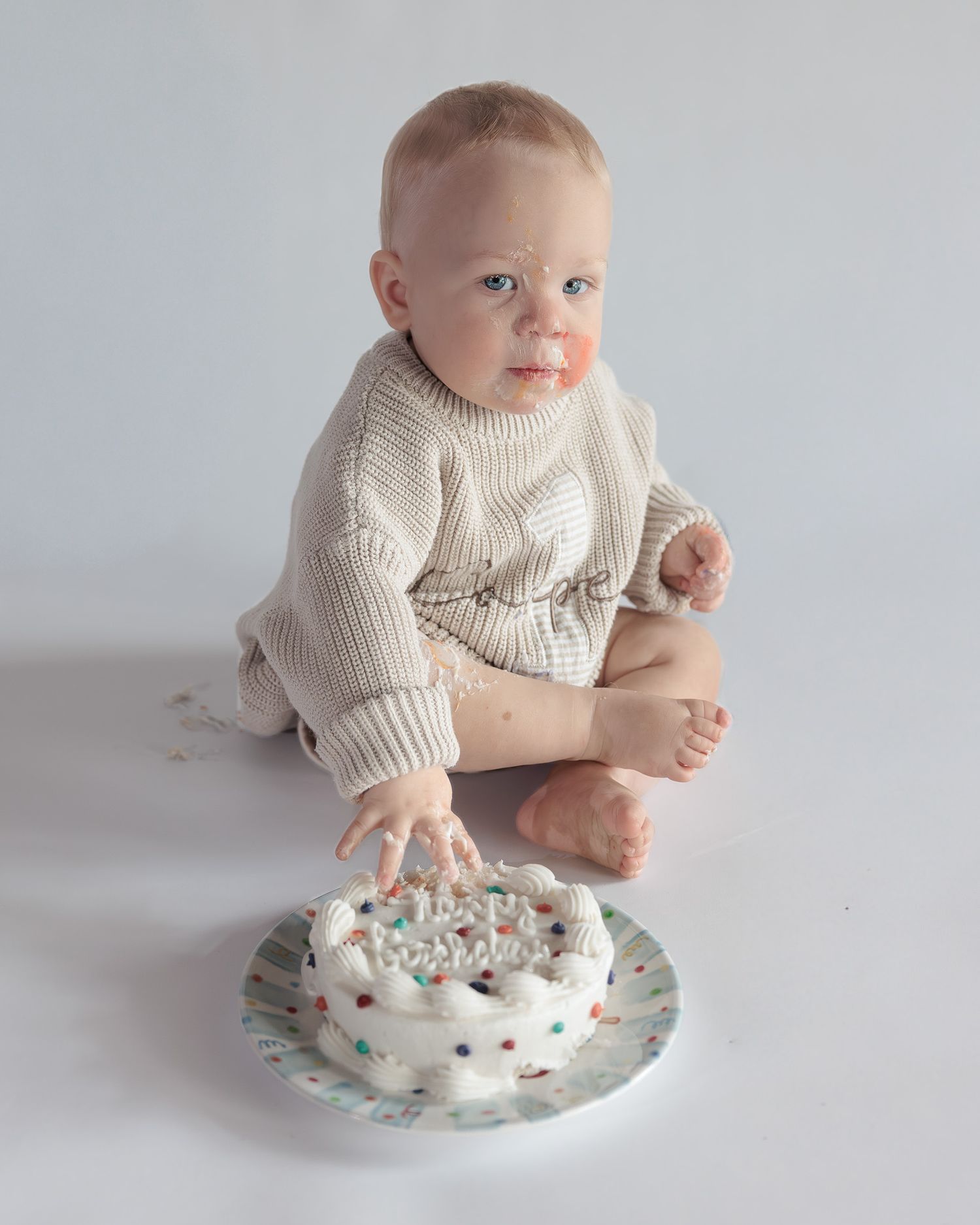 Baby boy in cream knit romper touches his white first birthday smash cake with colorful sprinkles on white background.