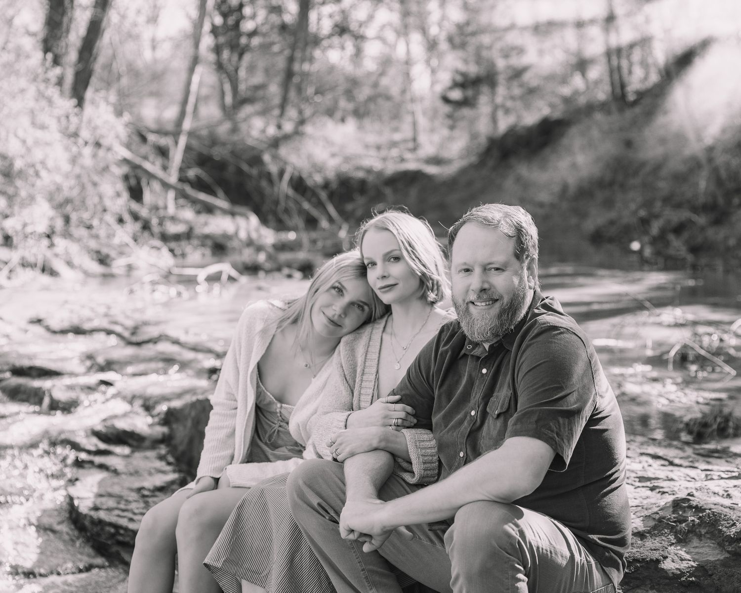 Black and white family portrait of two women and a bearded man sitting together by a creek in a wooded outdoor setting.