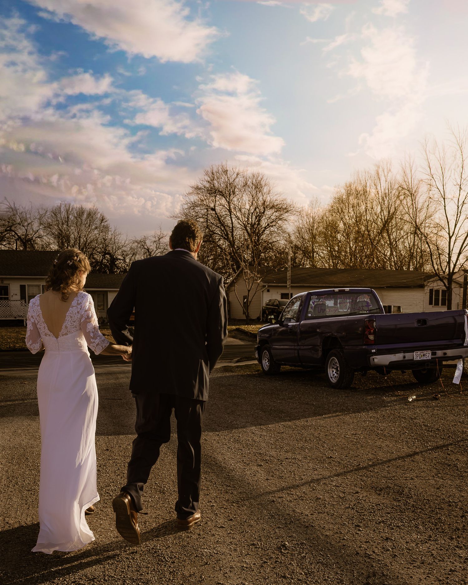 Outdoor wedding scene at sunset with silhouettes walking across gravel parking area near parked vehicles.