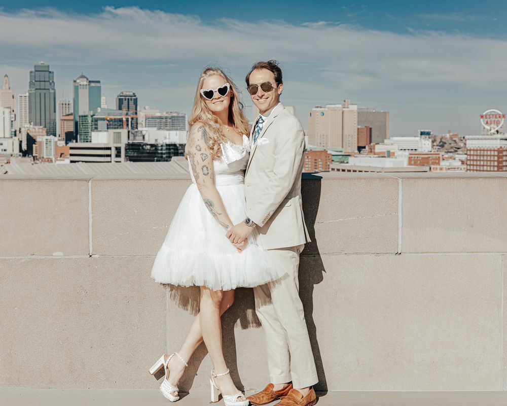 A couple poses together on a sunny rooftop with urban buildings in the background.