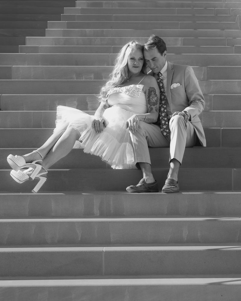 A romantic black and white photo of a couple sitting on outdoor steps, the bride in a white dress and heels.