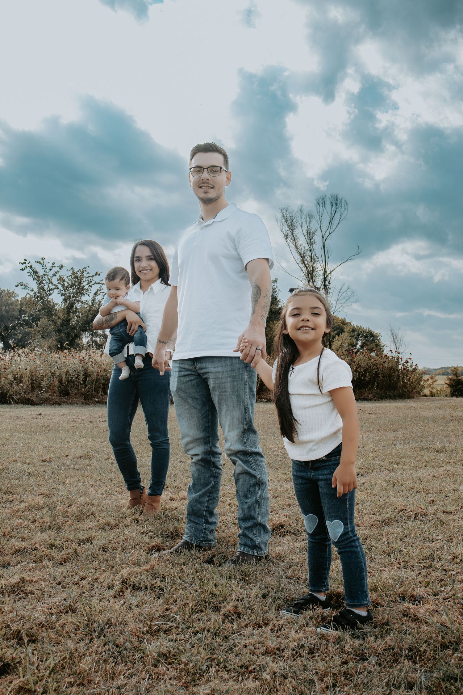 Family of four posing outdoors in a grassy field, parents and two young children wearing white tops and jeans under cloudy skies.