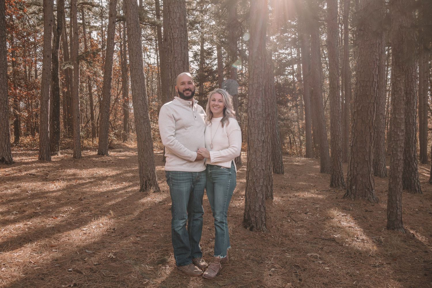 A couple stands together on a sunlit forest path surrounded by tall pine trees on an autumn afternoon.