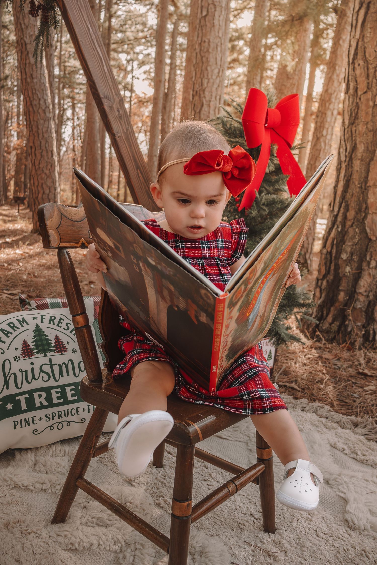 A child in red and plaid holiday attire sits in a rustic wooden chair next to a Christmas tree farm sign.