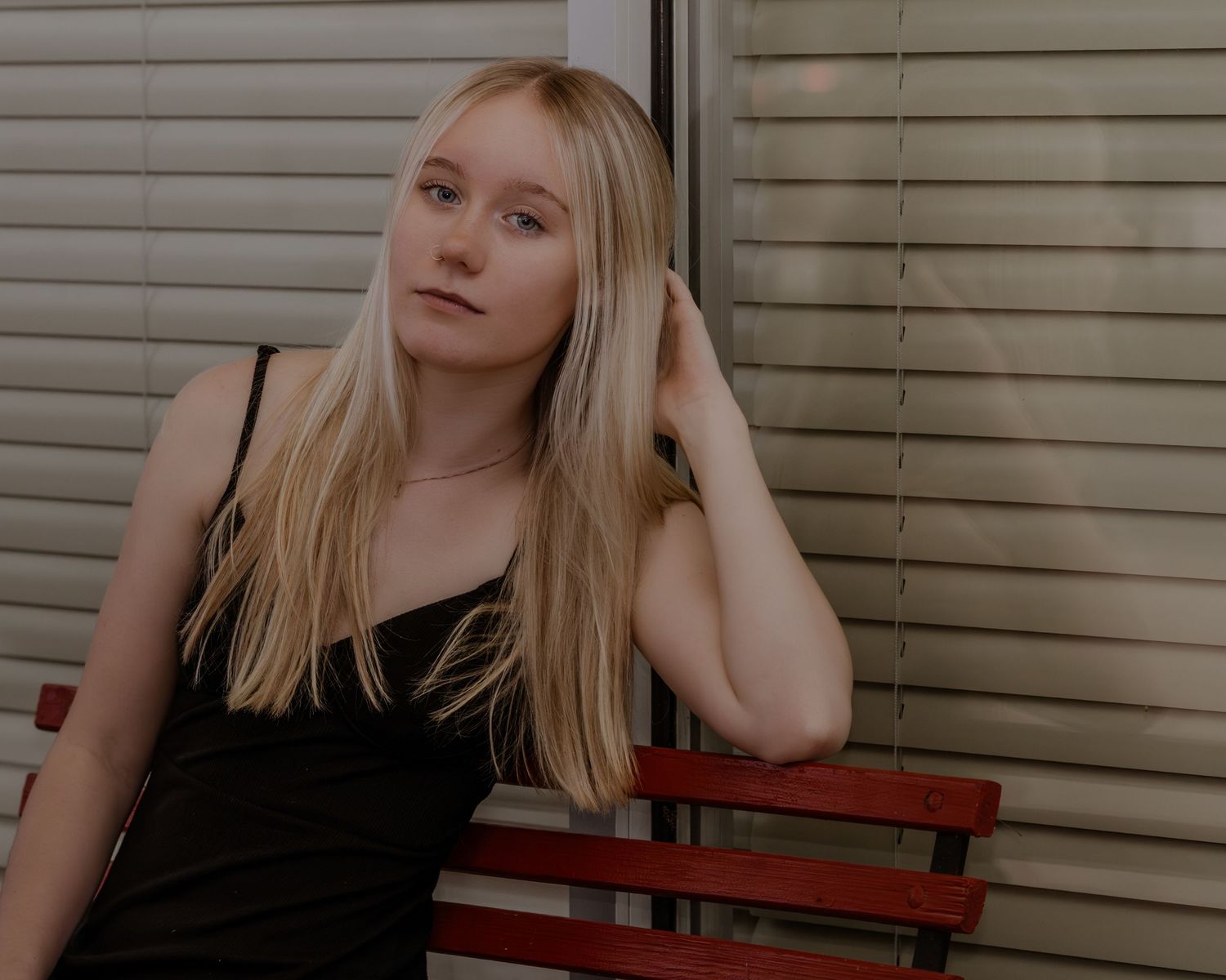 A person with blonde hair wearing a black top poses on a red bench against a white wall.