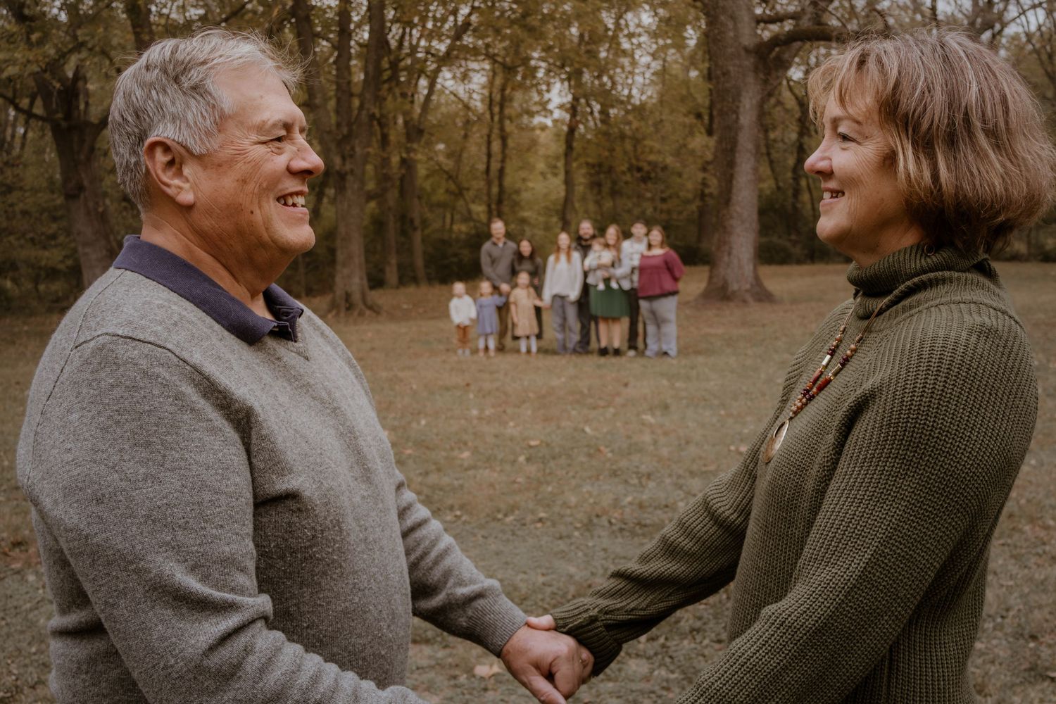 Older couple holding hands and smiling at each other with extended family gathered in autumn woodland background.