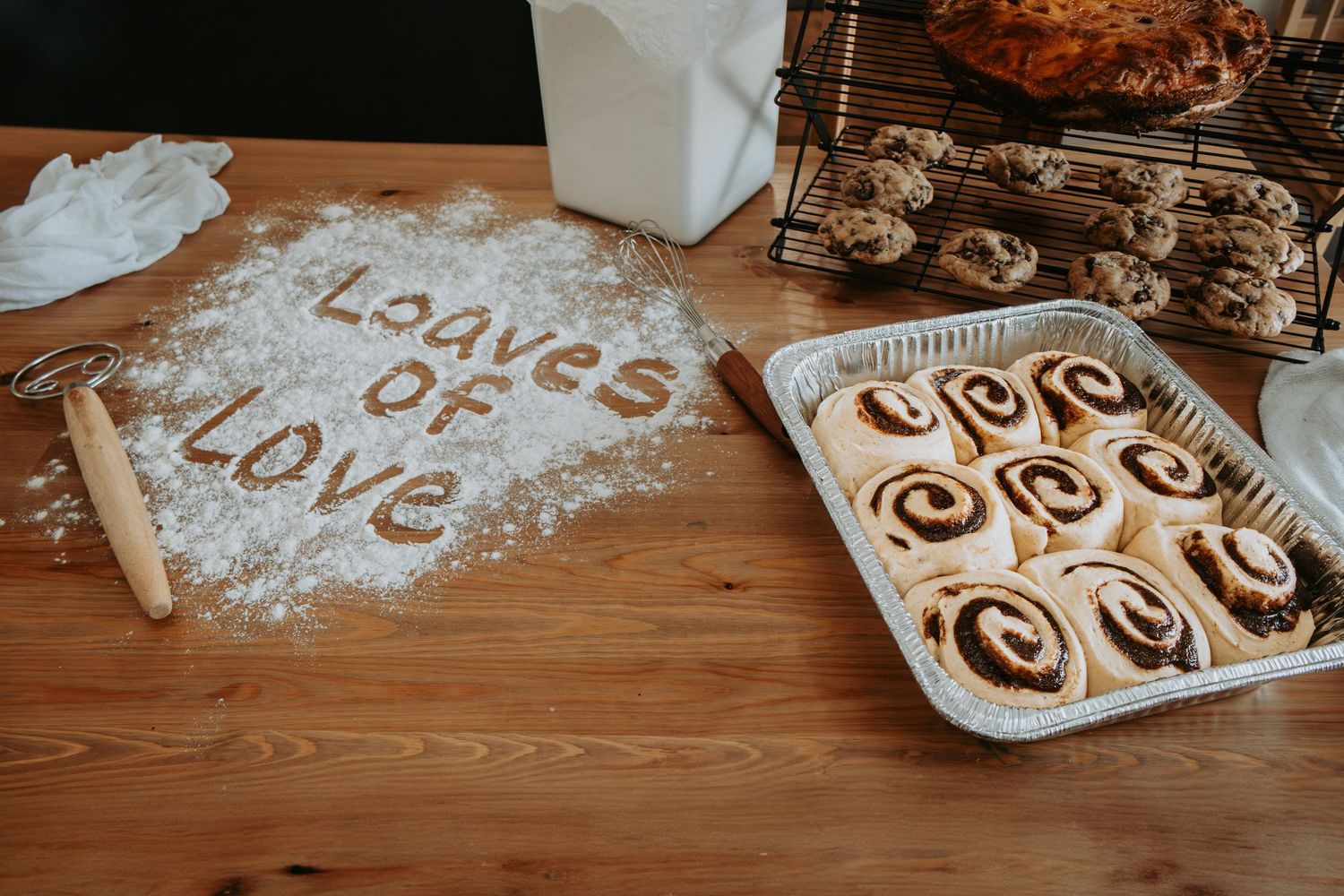 Cinnamon rolls in a foil pan beside flour-dusted table with Loaves of Love written in cinnamon, rolling pin, and cooling rack.
