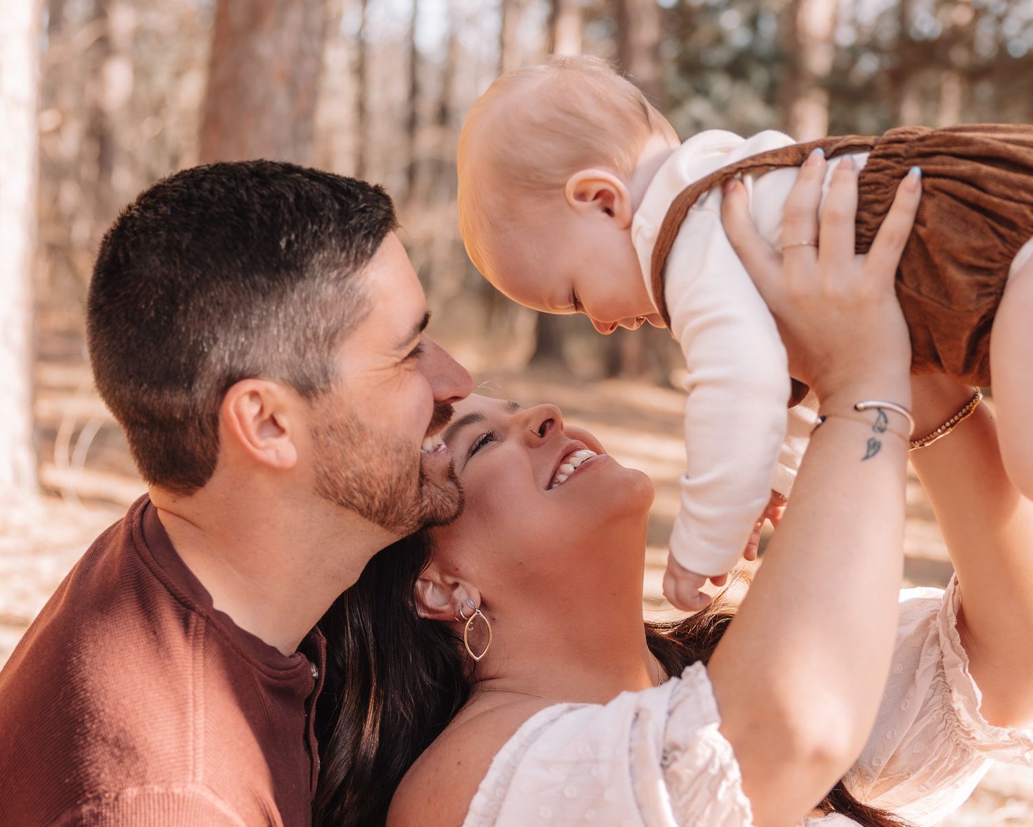 Happy couple holding smiling baby outdoors in sunlit forest during a joyful family photo session.