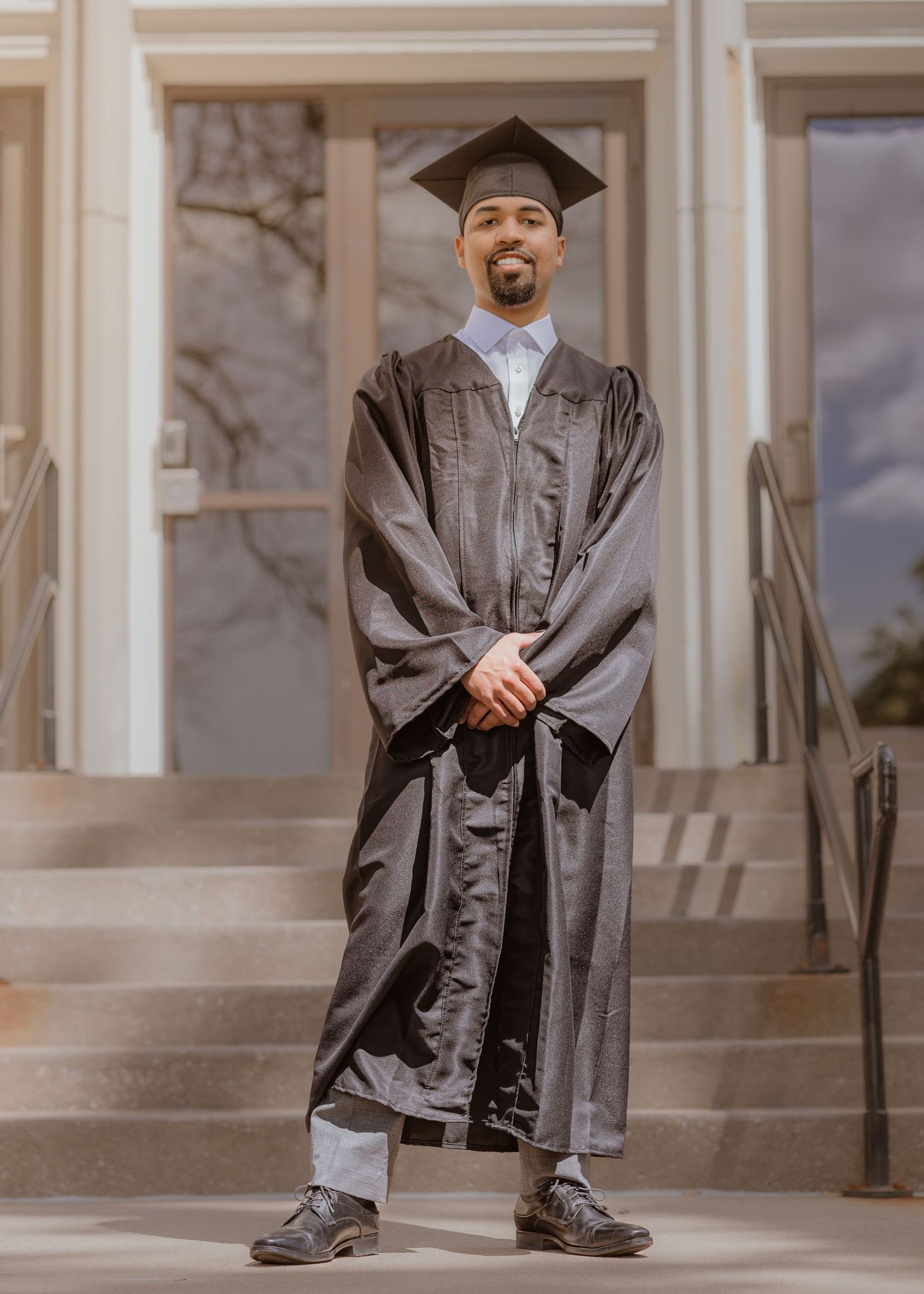 Graduate in black cap and gown smiling on university steps during outdoor graduation photo session.