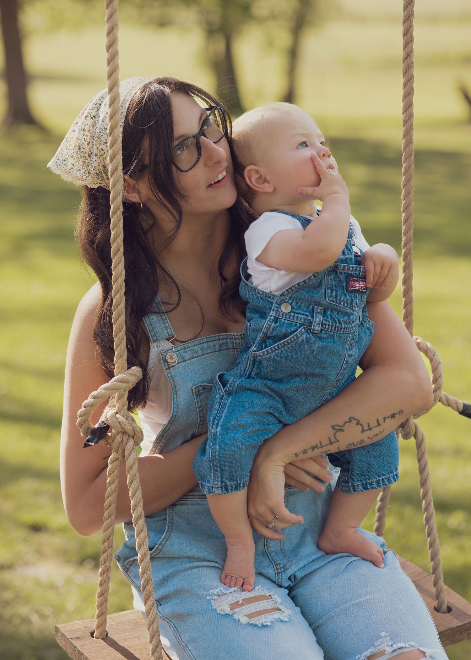 Young woman in denim overalls sitting on a rope swing holding a smiling baby outdoors in a sunny park.