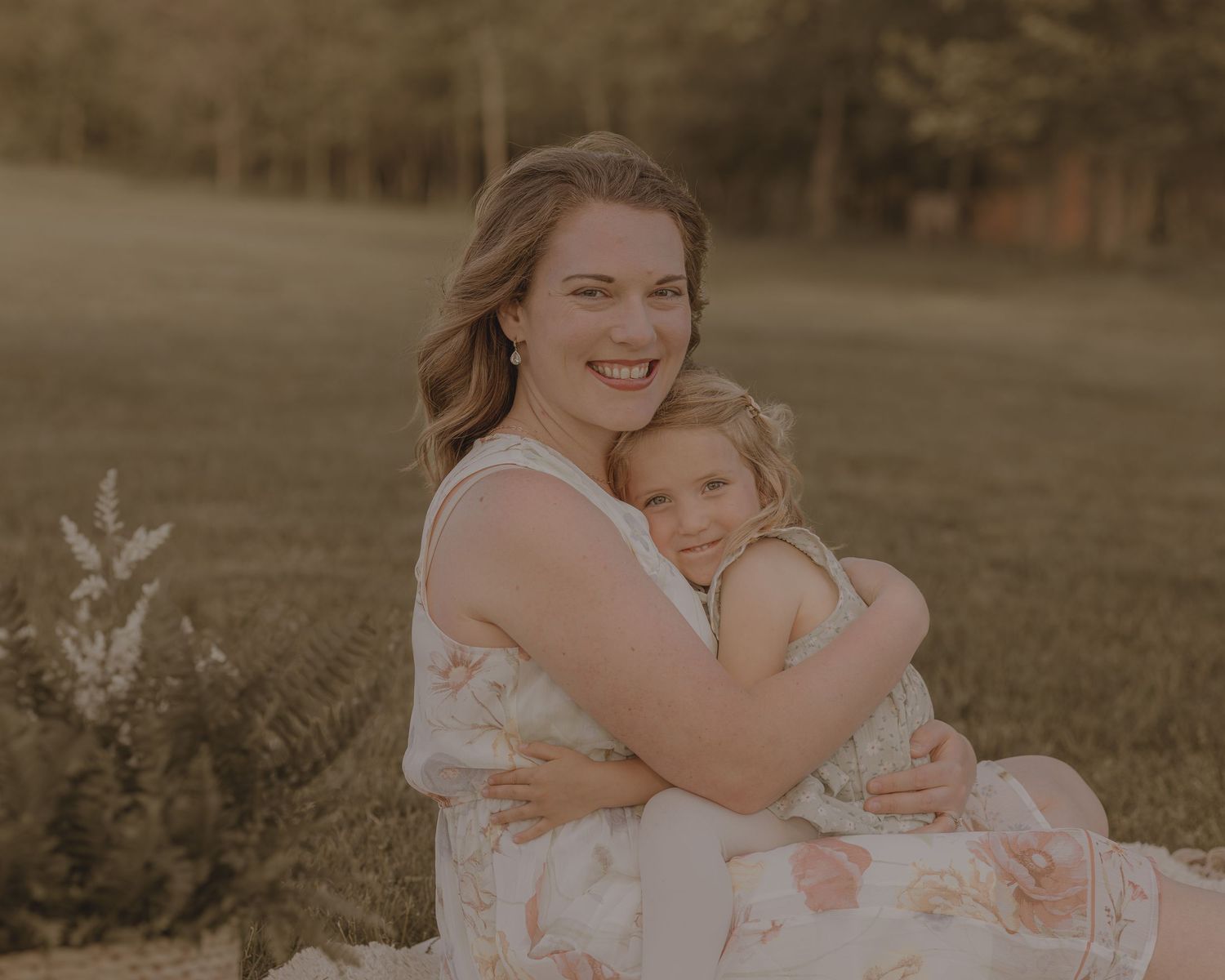 A smiling mother hugs her young daughter close while sitting outdoors in warm golden sunlight.