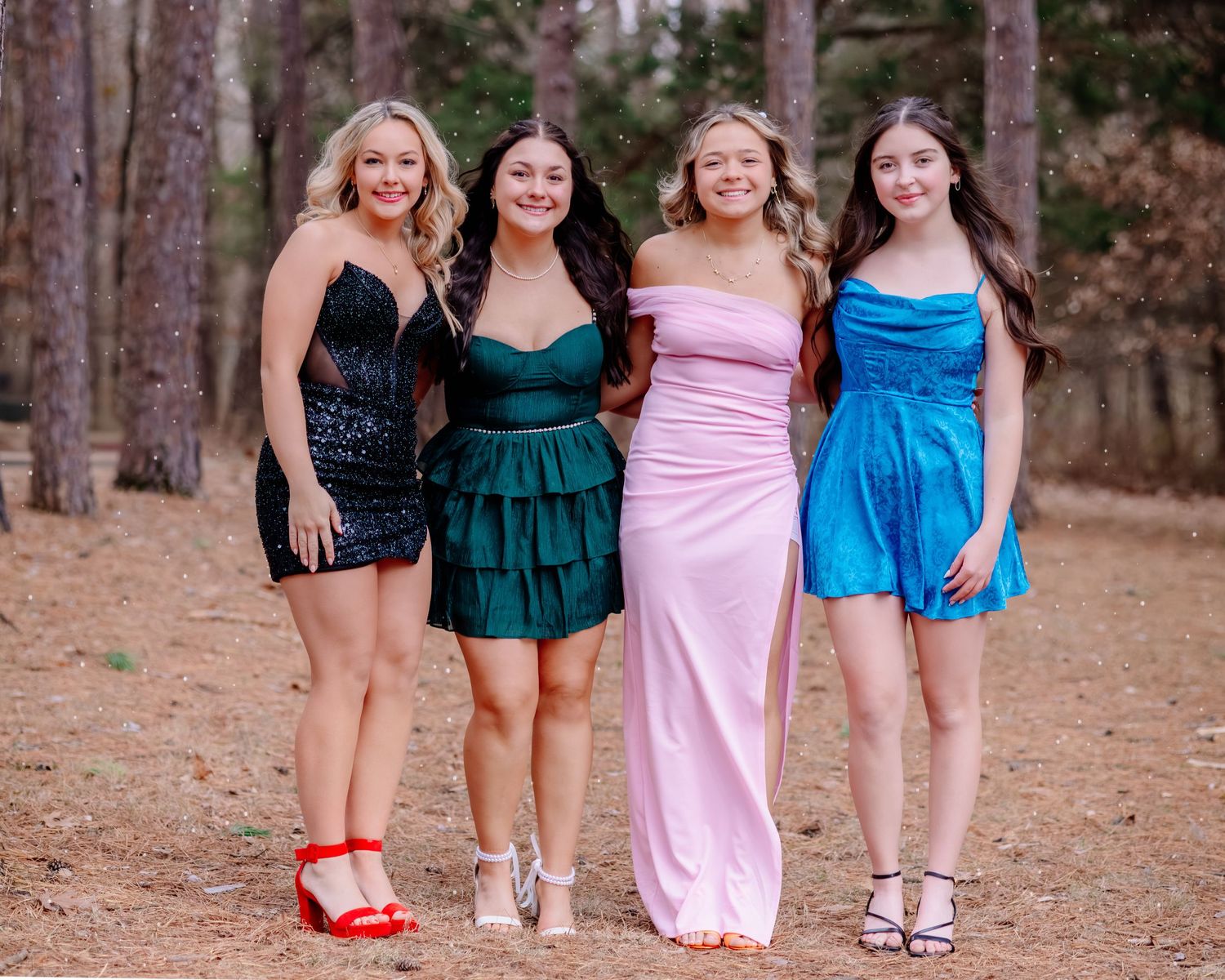 Four friends in elegant dresses pose together on a forest path, wearing black, green, pink and blue gowns.
