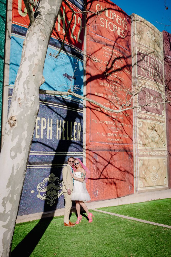 A figure in a white dress and pink shoes poses against a colorful mural wall with book spines.