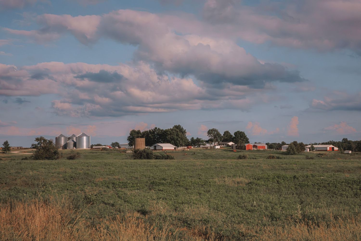 A rural farm with grain silos and red barns surrounded by green fields under a partly cloudy sky.