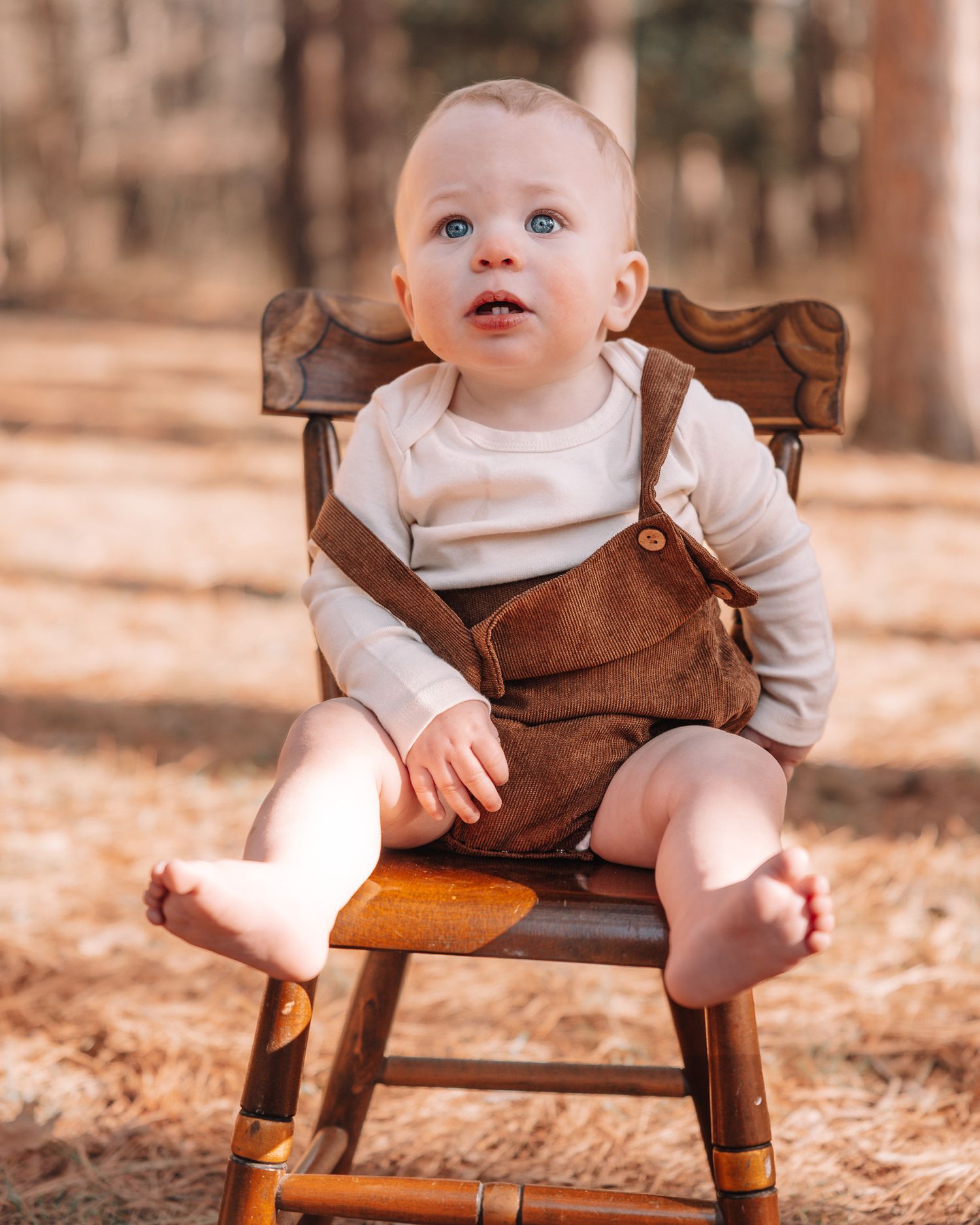 Baby wearing brown overalls sits on wooden chair in forest setting.