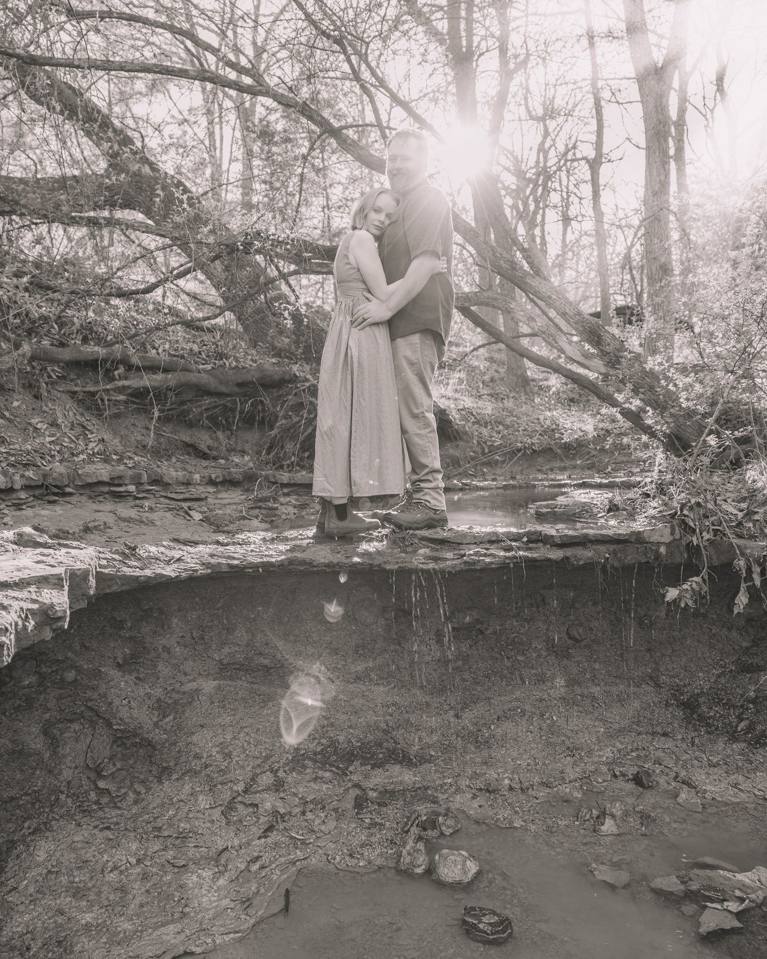Black and white photo of a couple embracing in a sunlit forest near a creek with fallen trees.