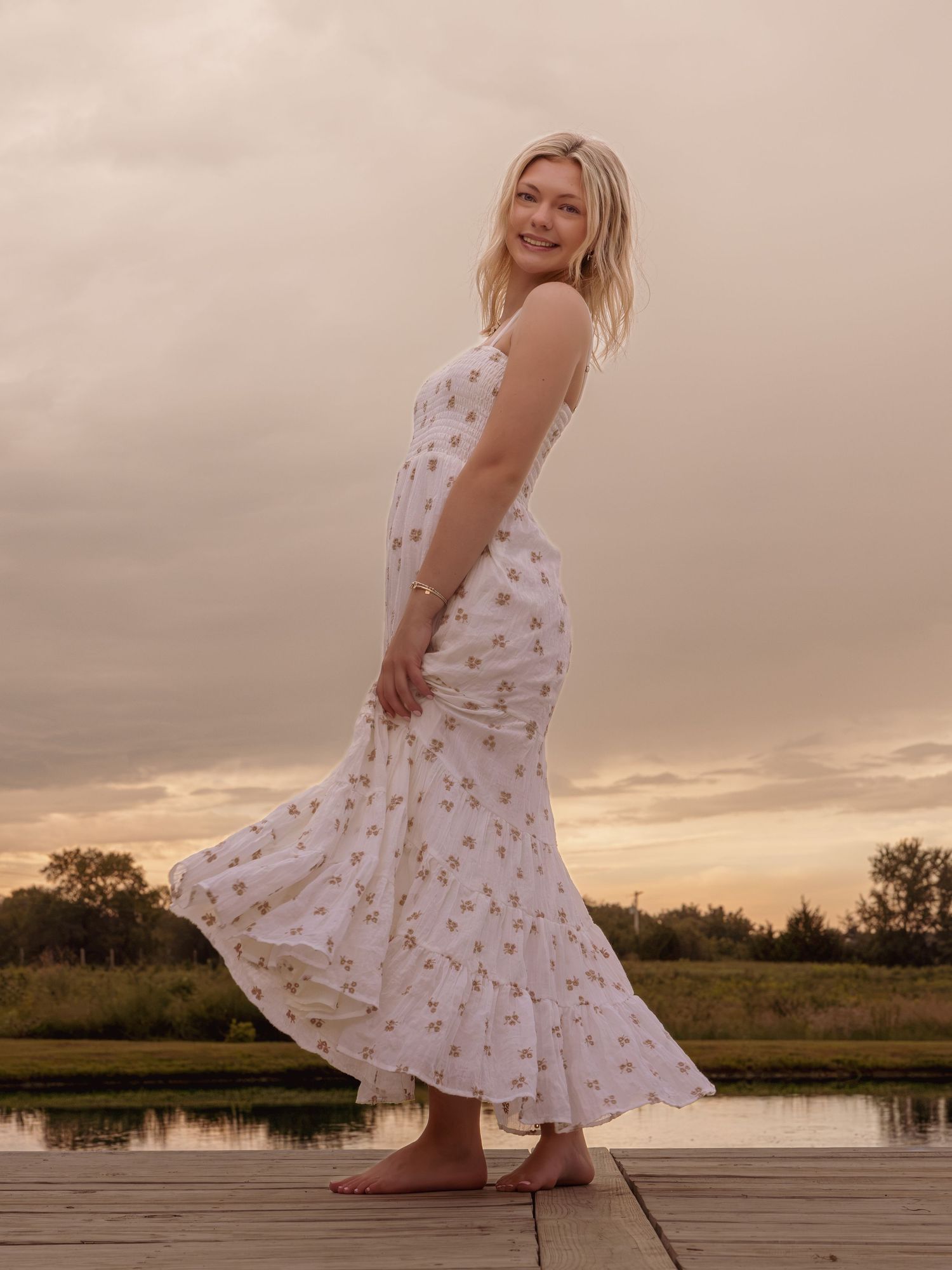 Blonde woman in floral white maxi dress posing on dock at sunset by a calm lake.