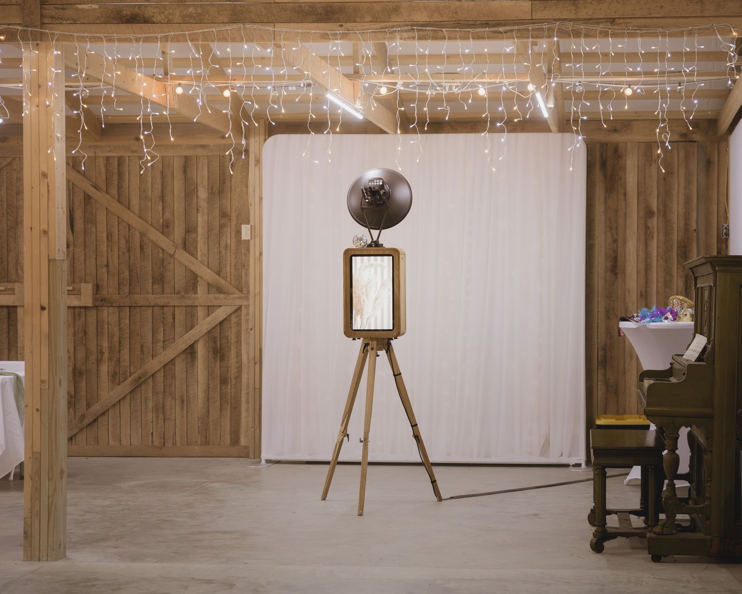 Rustic barn venue with fairy lights, white backdrop, vintage photo booth on tripod, and antique piano for wedding reception.