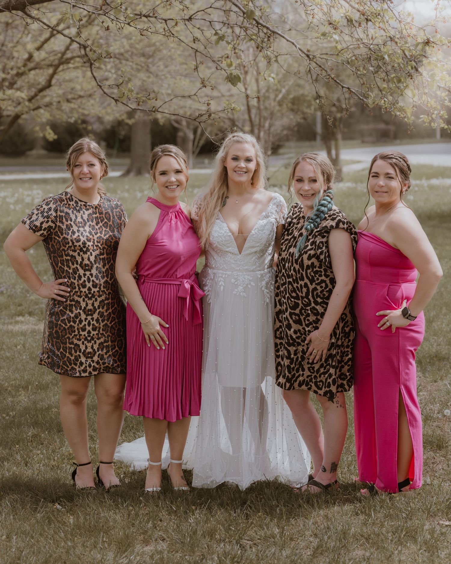 Bride in white gown poses with four bridesmaids in pink and leopard print dresses outdoors.