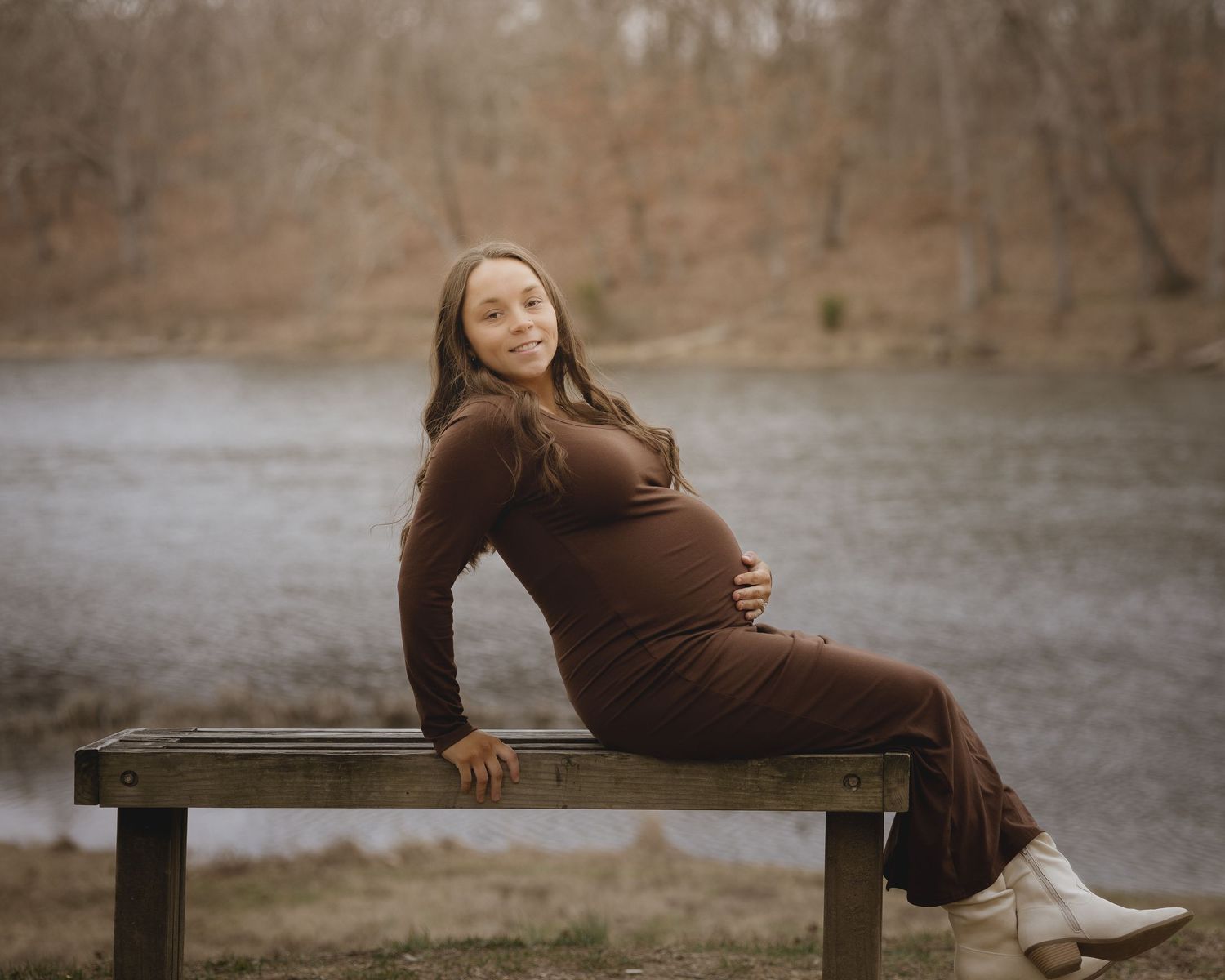 A pregnant woman in a brown dress poses on a wooden bench during an outdoor maternity photoshoot.
