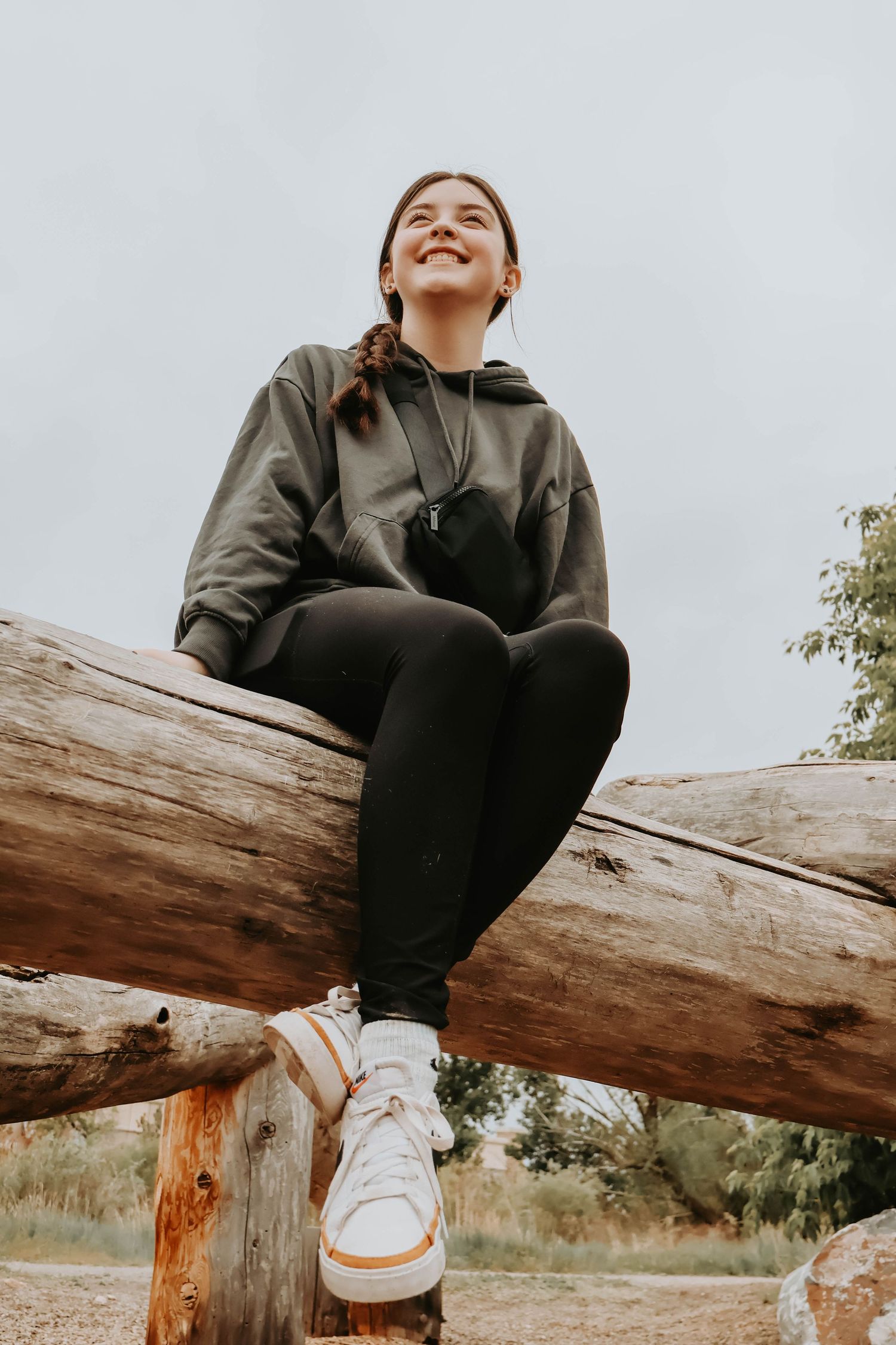 Person wearing black hoodie and sneakers sits joyfully on wooden log in outdoor setting during daytime.