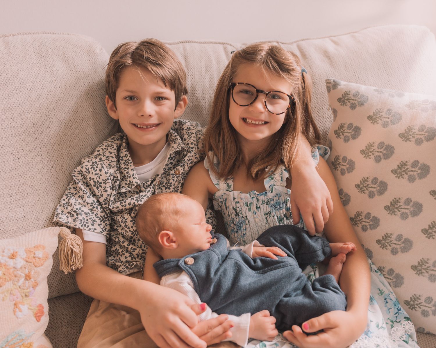 Two children and a newborn baby sibling pose together on a couch, smiling warmly in a cozy home setting.