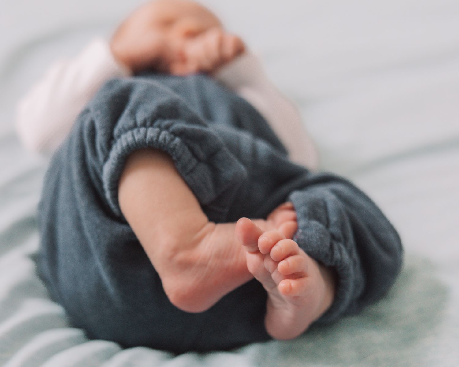 Close-up of a sleeping newborn baby's tiny feet in soft blue linen pants on a light blanket.