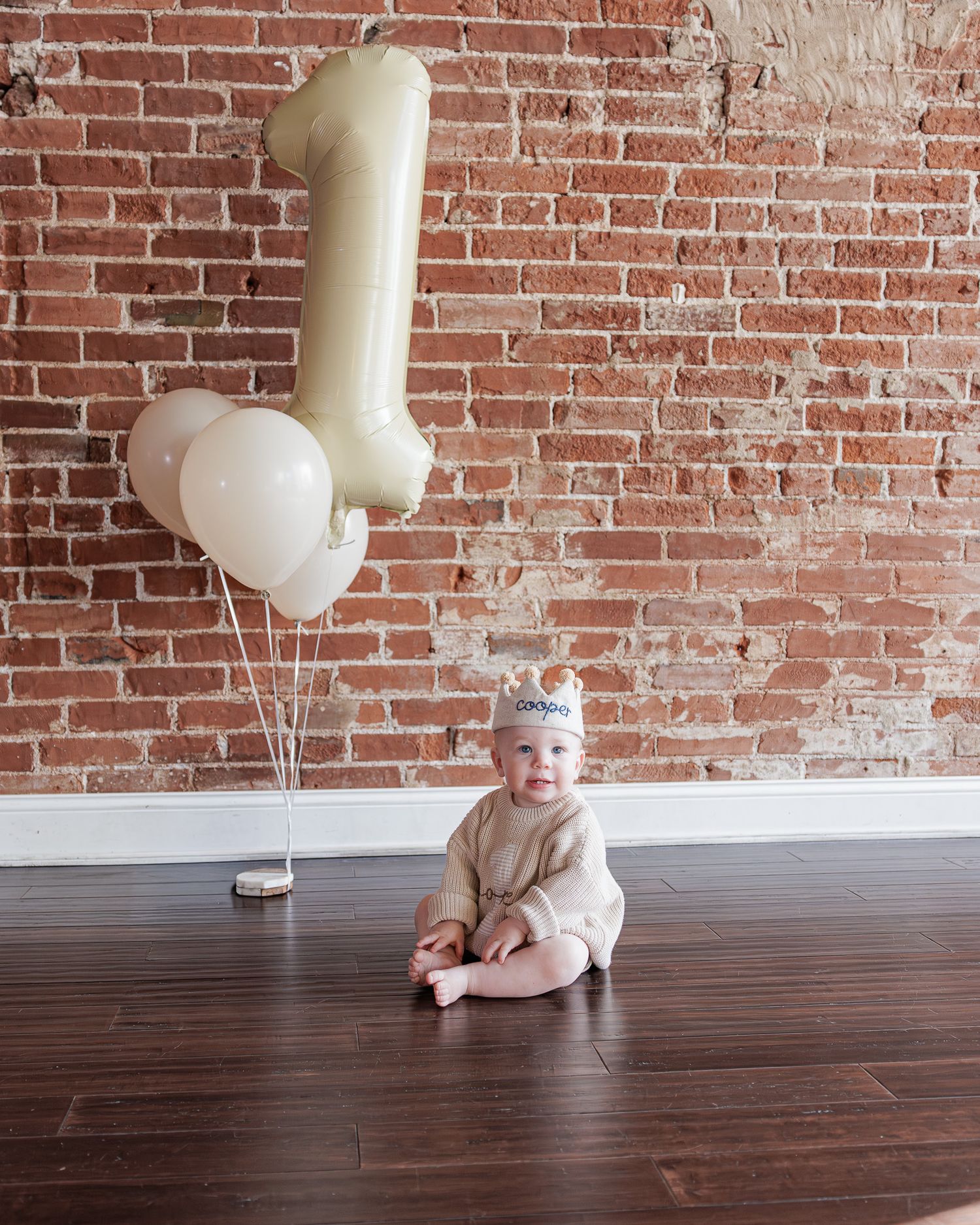 Adorable baby celebrates first birthday sitting before number one balloons against brick wall.