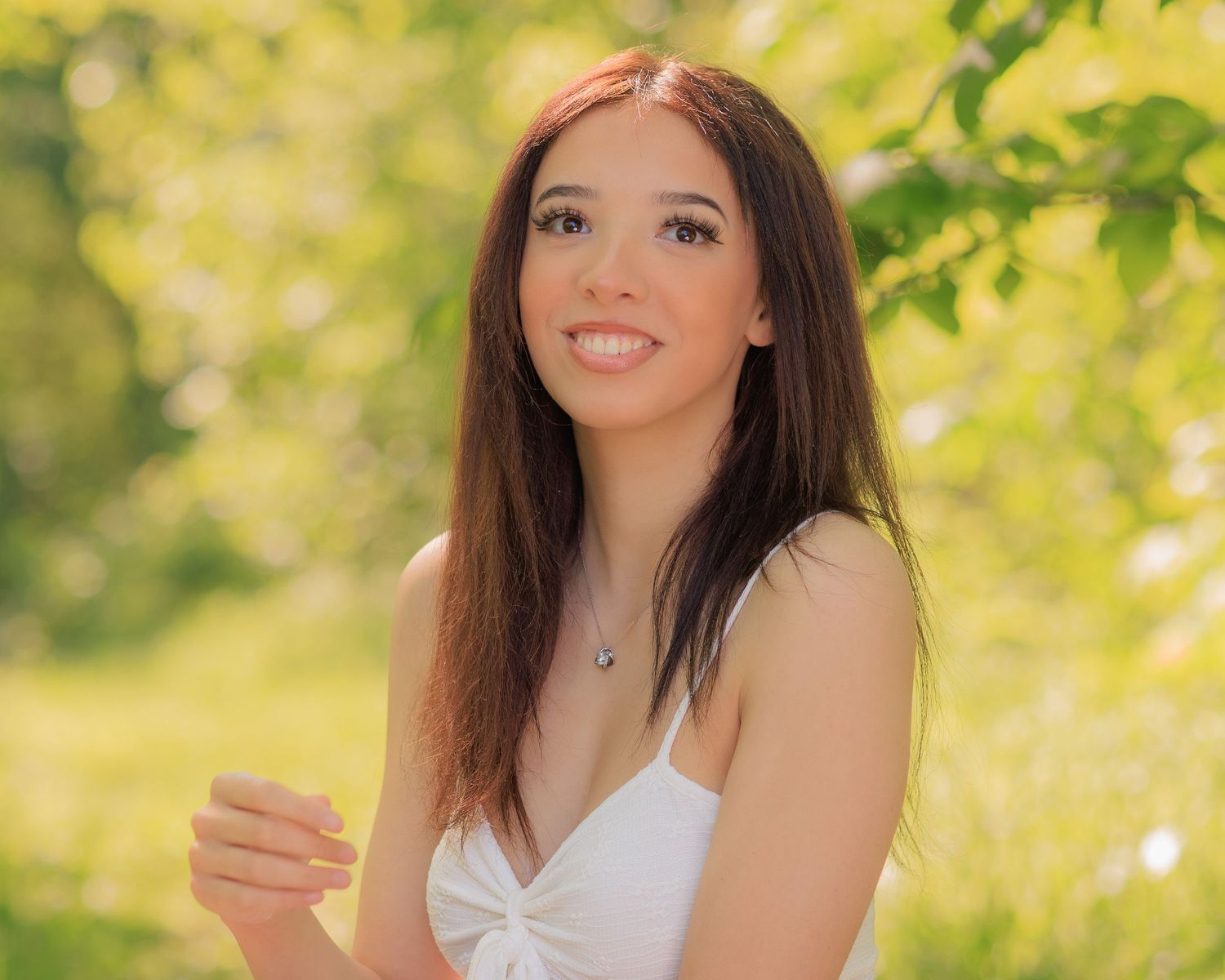 Young woman with long brown hair smiling outdoors in a white dress, surrounded by lush green foliage.