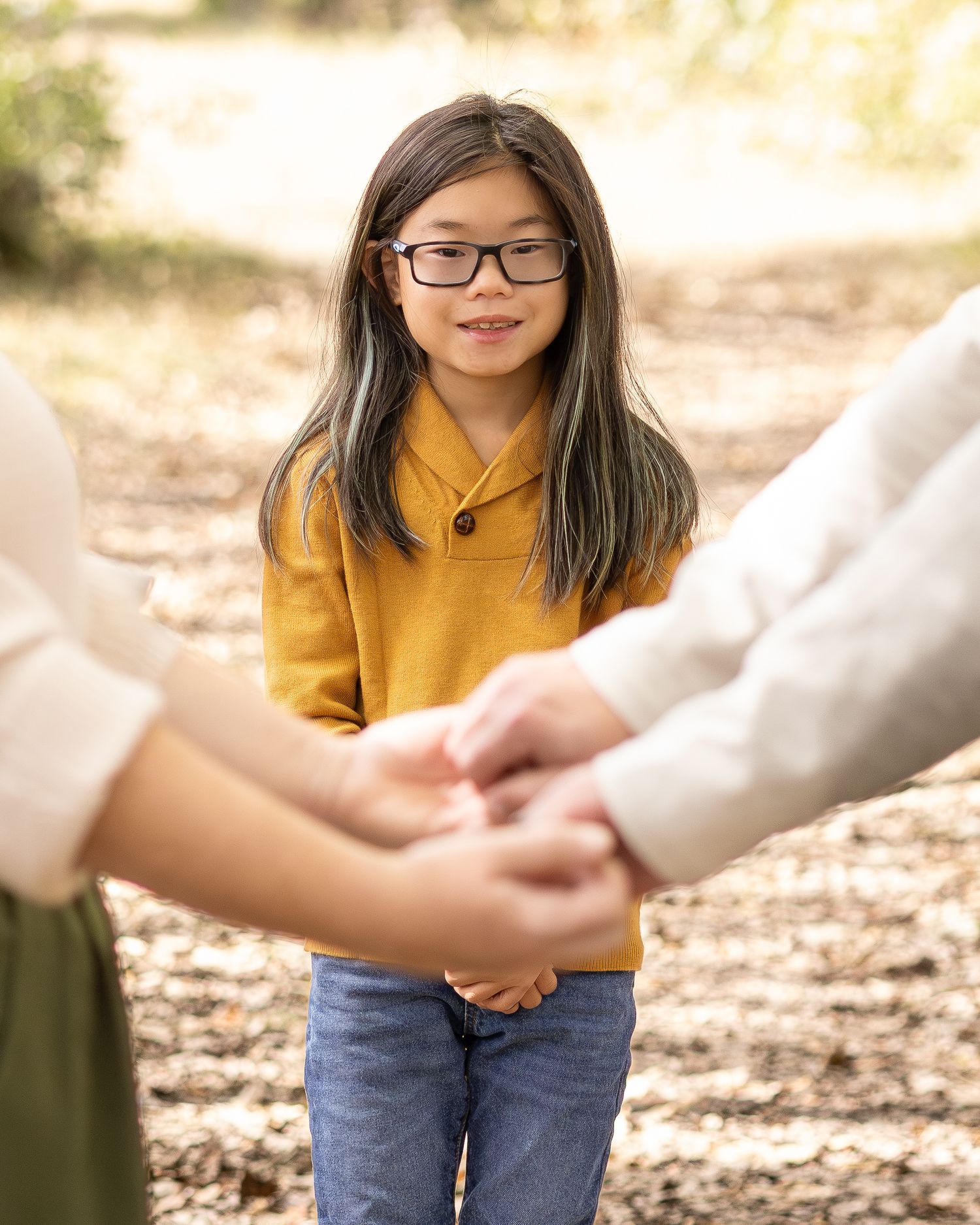 A young Asian girl with glasses and a yellow sweater smiles warmly while holding hands with adults outdoors.