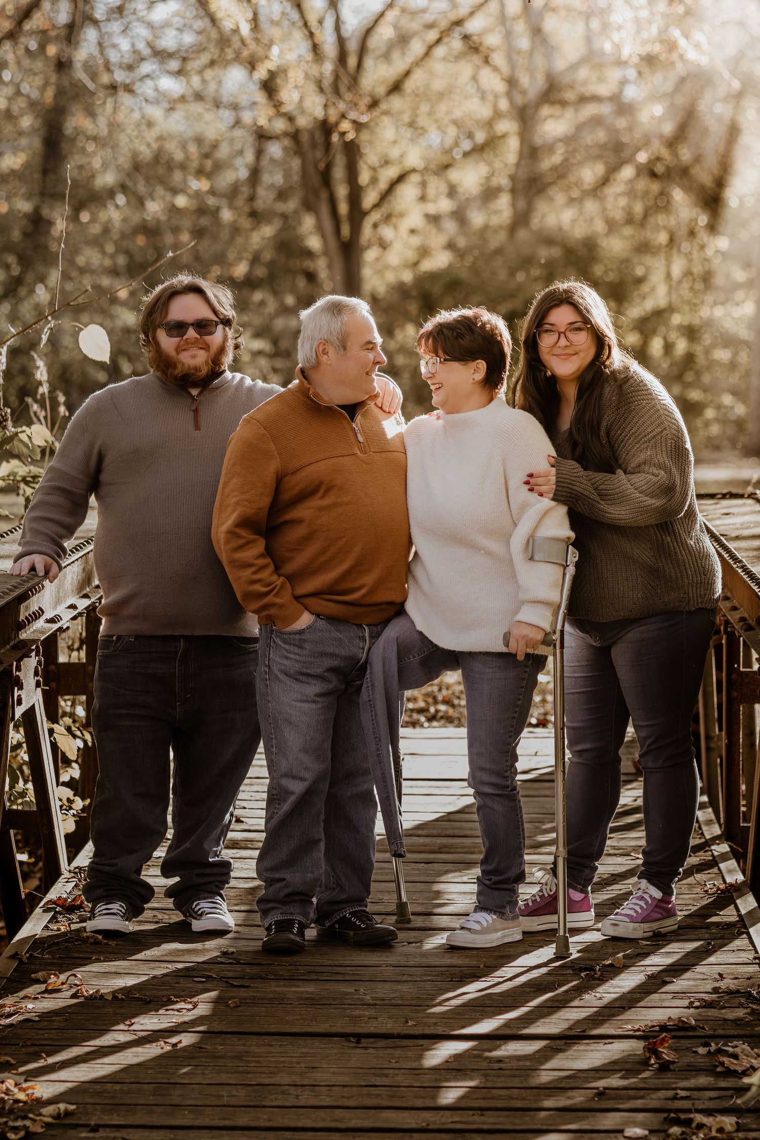 A group in cozy sweaters stands together on a wooden bridge during golden hour in an autumn setting.