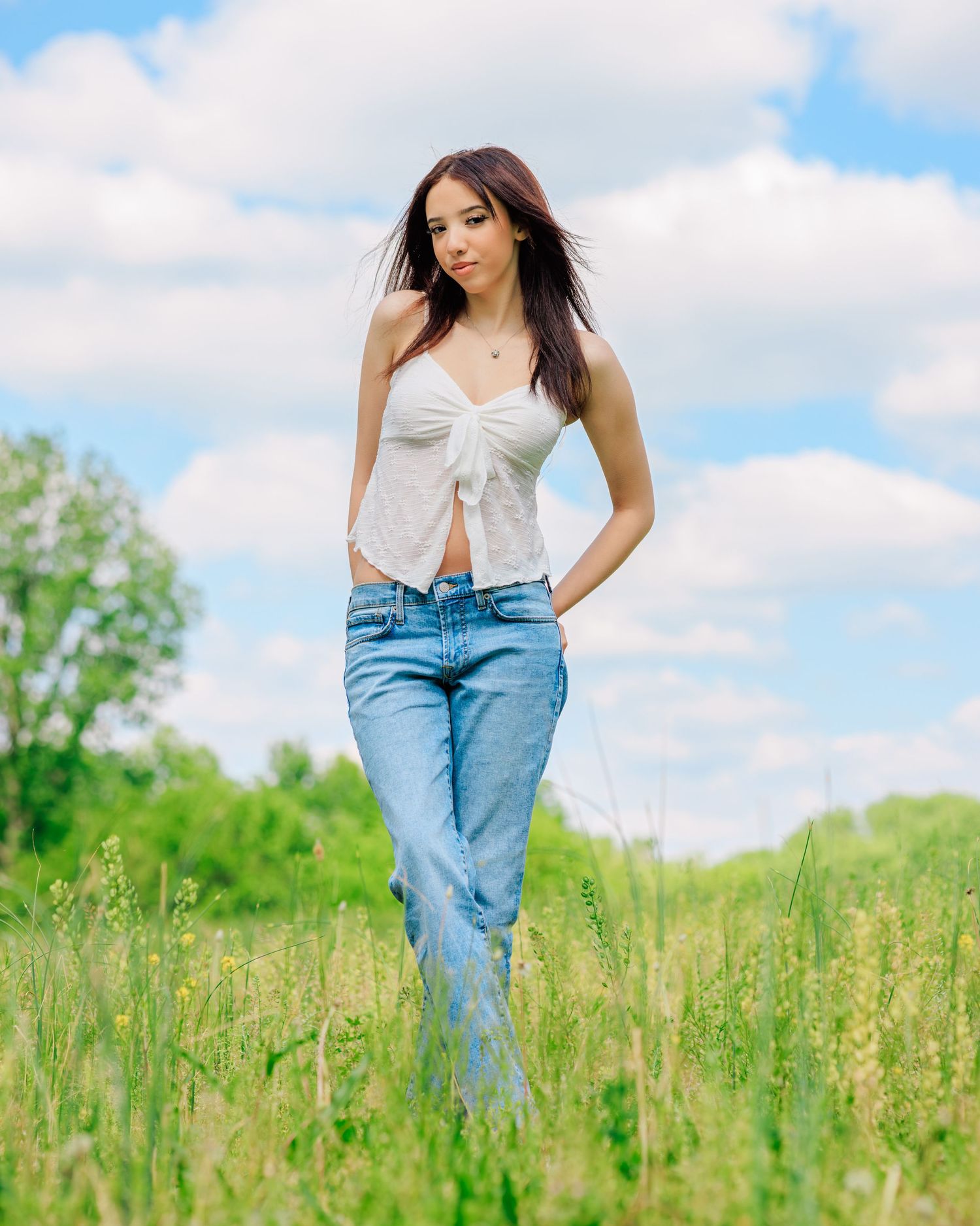 Young woman in white crop top and blue jeans standing in a green meadow under a cloudy sky.