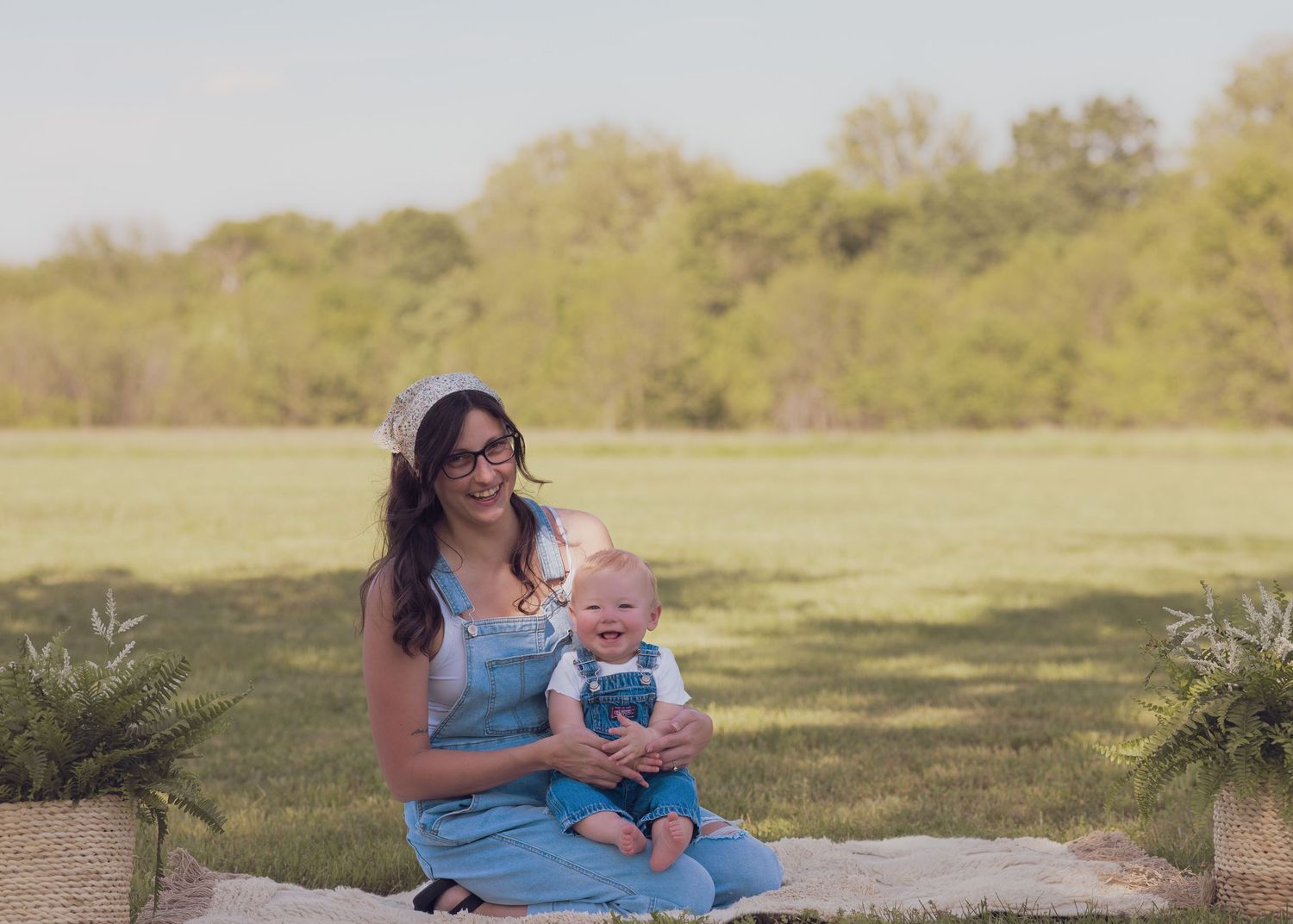 Mother in denim overalls smiling with baby boy outdoors on blanket in green field.