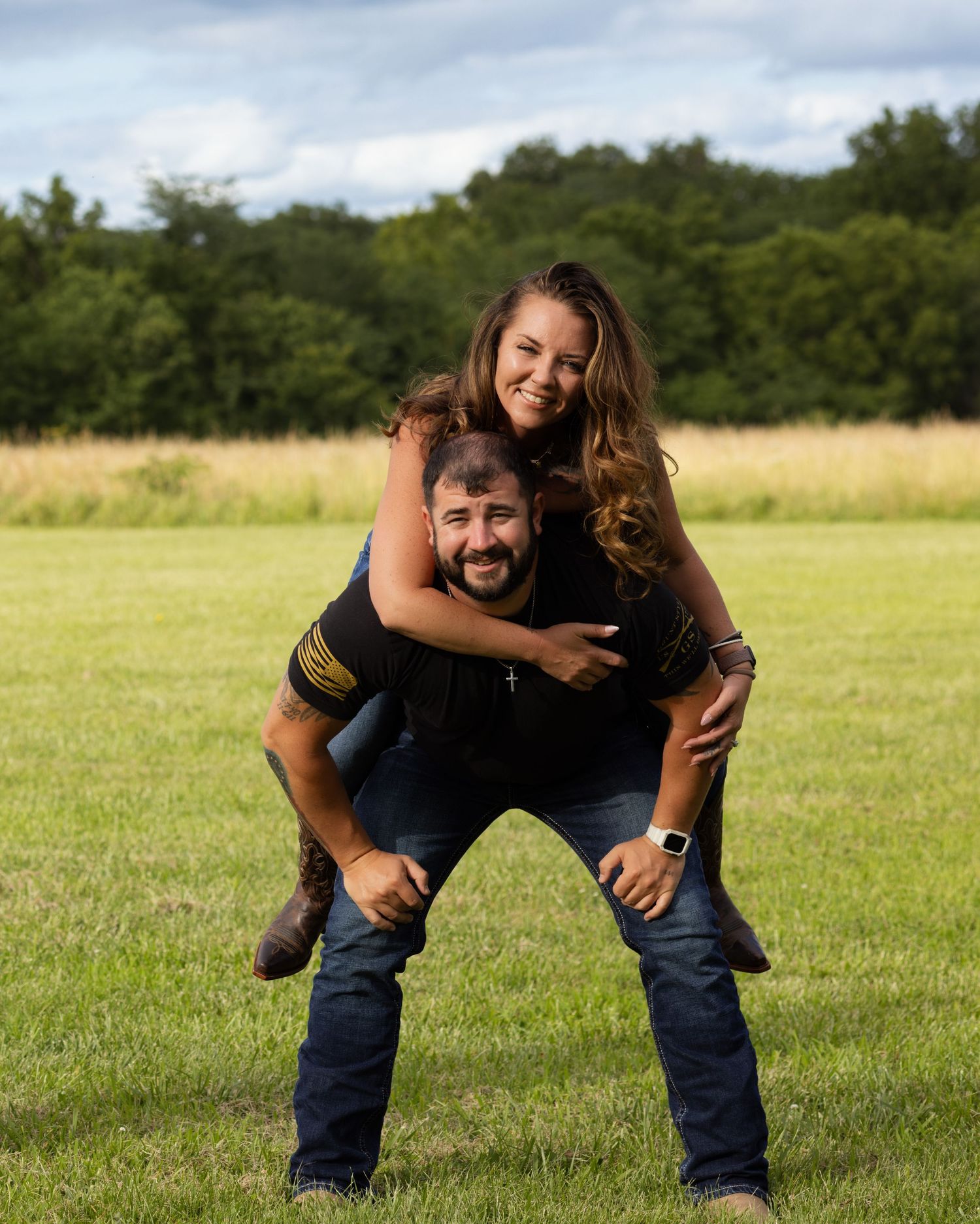 Happy couple posing playfully in green field, woman riding piggyback on mans back outdoors.