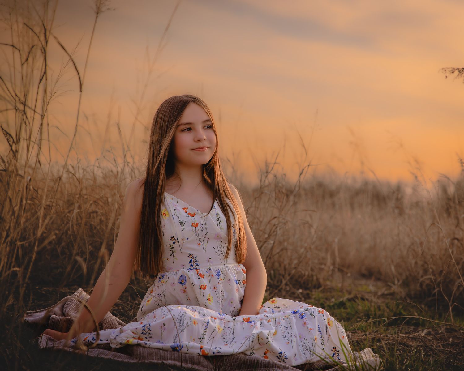 A person in a floral dress sits in a golden field at sunset creating a dreamy summer portrait.