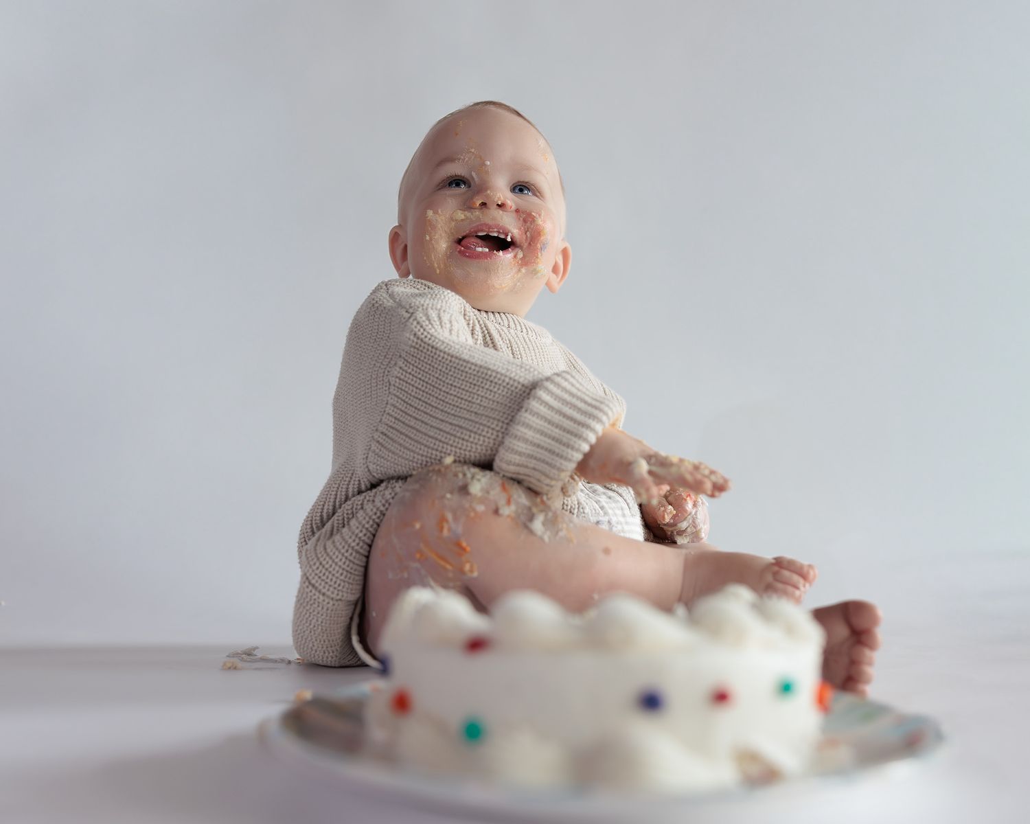 Smiling baby in white knit outfit celebrating first birthday with a white smash cake covered in colorful sprinkles.