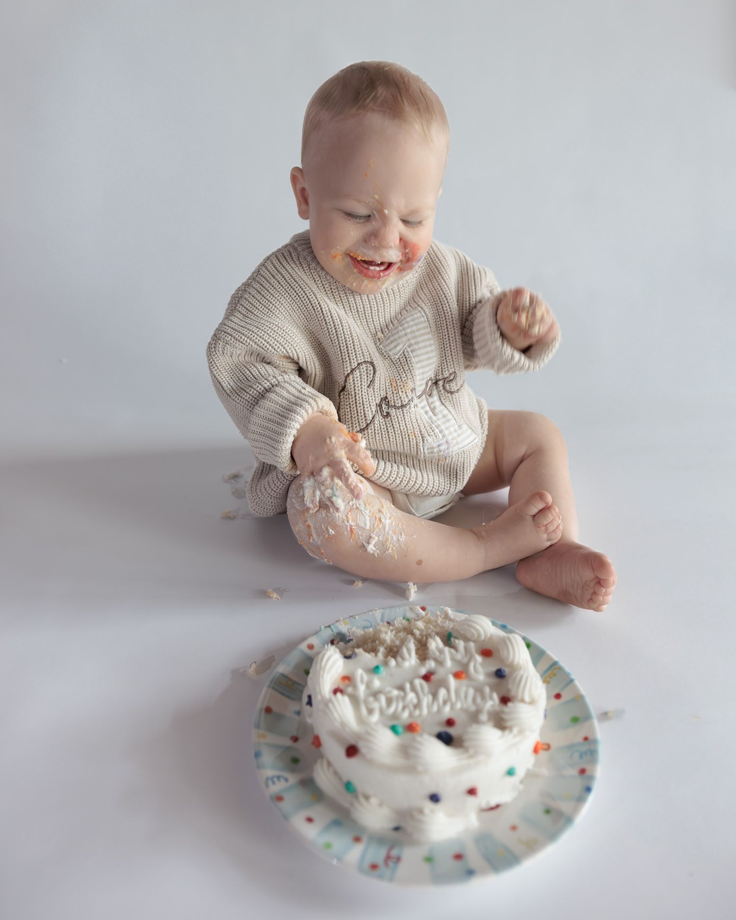 Happy baby in a knit sweater smashes a white sprinkle cake during a first birthday cake smash photo session.