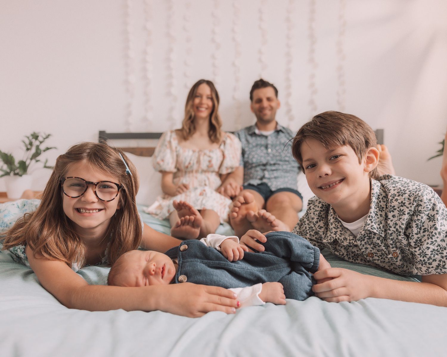 Family of five with two kids and newborn baby posing together on a bed in a bright white room.