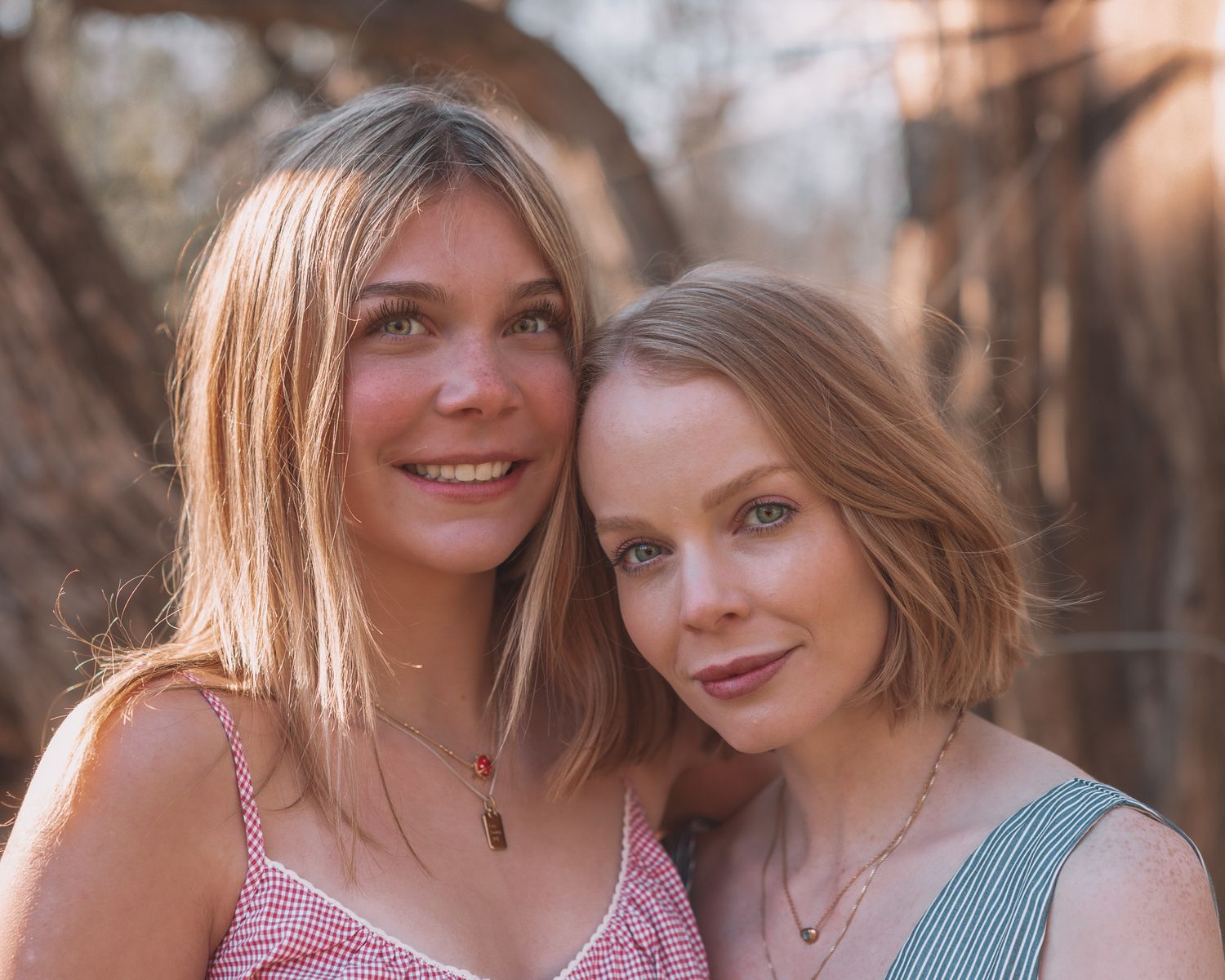 Two young blonde women smiling closely together outdoors in warm sunlight.